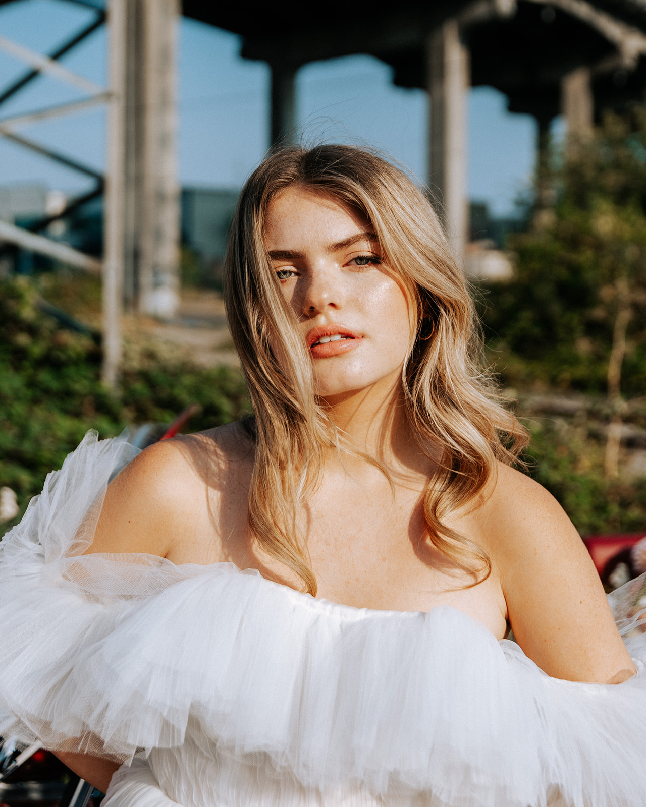 Carefree bad-ass bride wearing flirty Zephyr dress by local designer Ouma Bridal, standing in front of vintage red muscle car surrounded by tangerine and white summer blooms. 