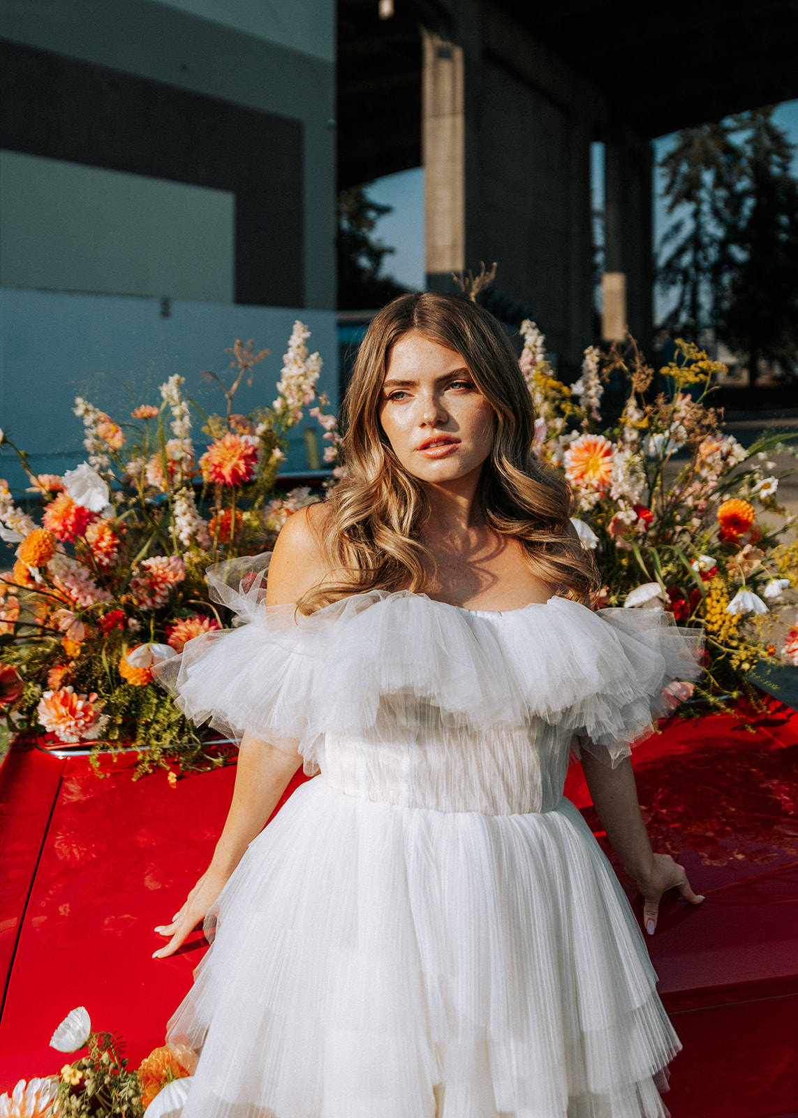 Carefree bad-ass bride wearing flirty Zephyr dress by local designer Ouma Bridal, standing in front of vintage red muscle car surrounded by tangerine and white summer blooms. 