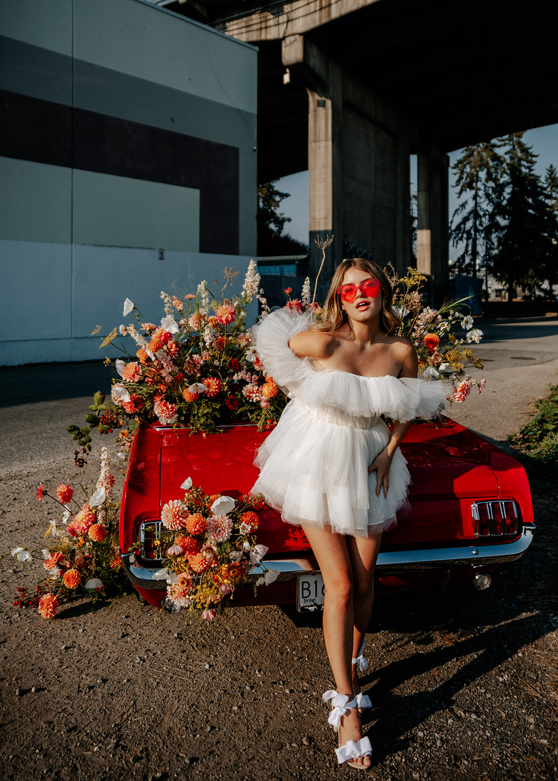 Carefree bad-ass bride wearing flirty Zephyr dress by local designer Ouma Bridal, and orange heart sunglasses, standing in front of vintage red muscle car surrounded by tangerine and white summer blooms. 