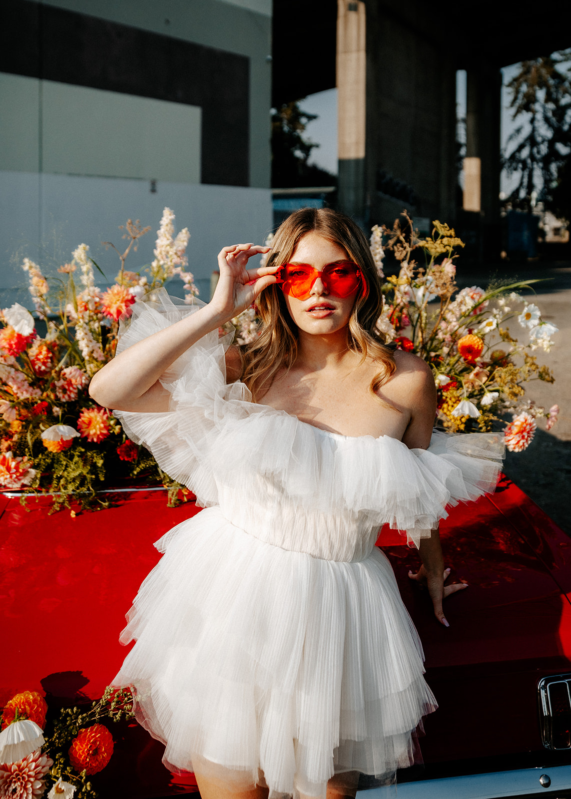 Carefree bad-ass bride wearing flirty Zephyr dress by local designer Ouma Bridal, and orange heart sunglasses, standing in front of vintage red muscle car surrounded by tangerine and white summer blooms. 