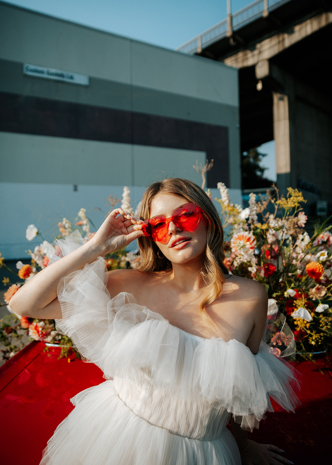 Bold bride wearing flirty dress and red heart sunglasses, standing in front of vintage red muscle car surrounded by summer blooms. 