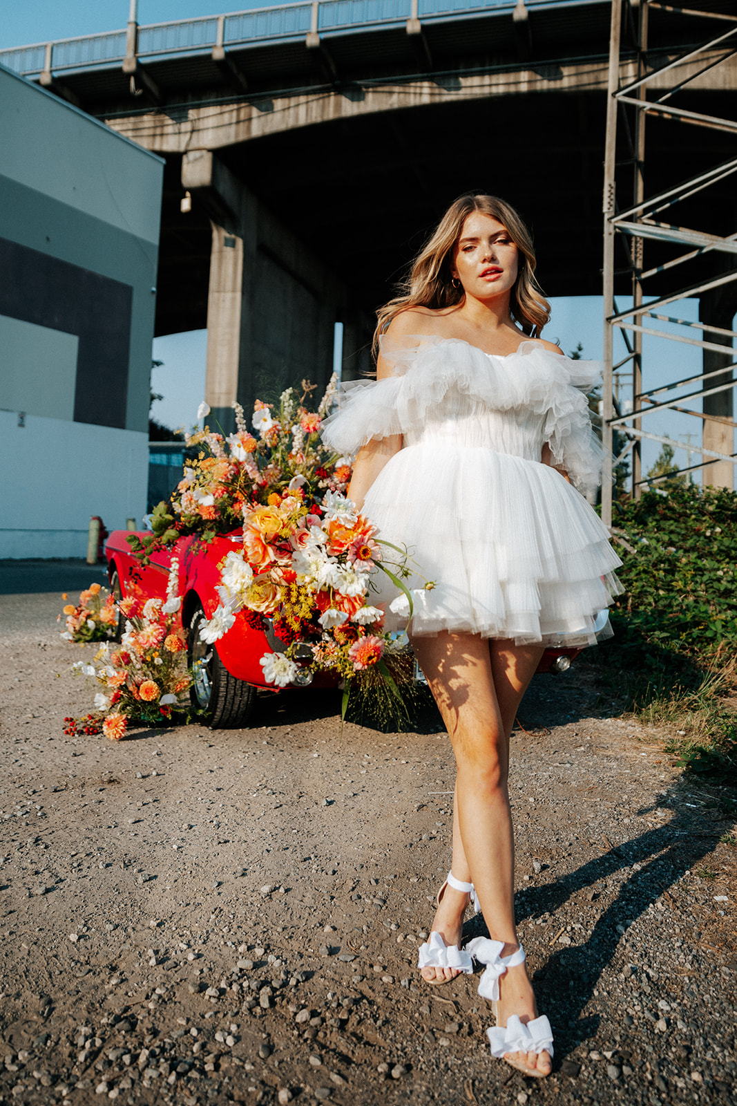 Carefree bad-ass bride wearing flirty Zephyr dress by local Canadian designer Ouma Bridal, standing in front of vintage red muscle car surrounded by tangerine, orange, and white summer flowers. 