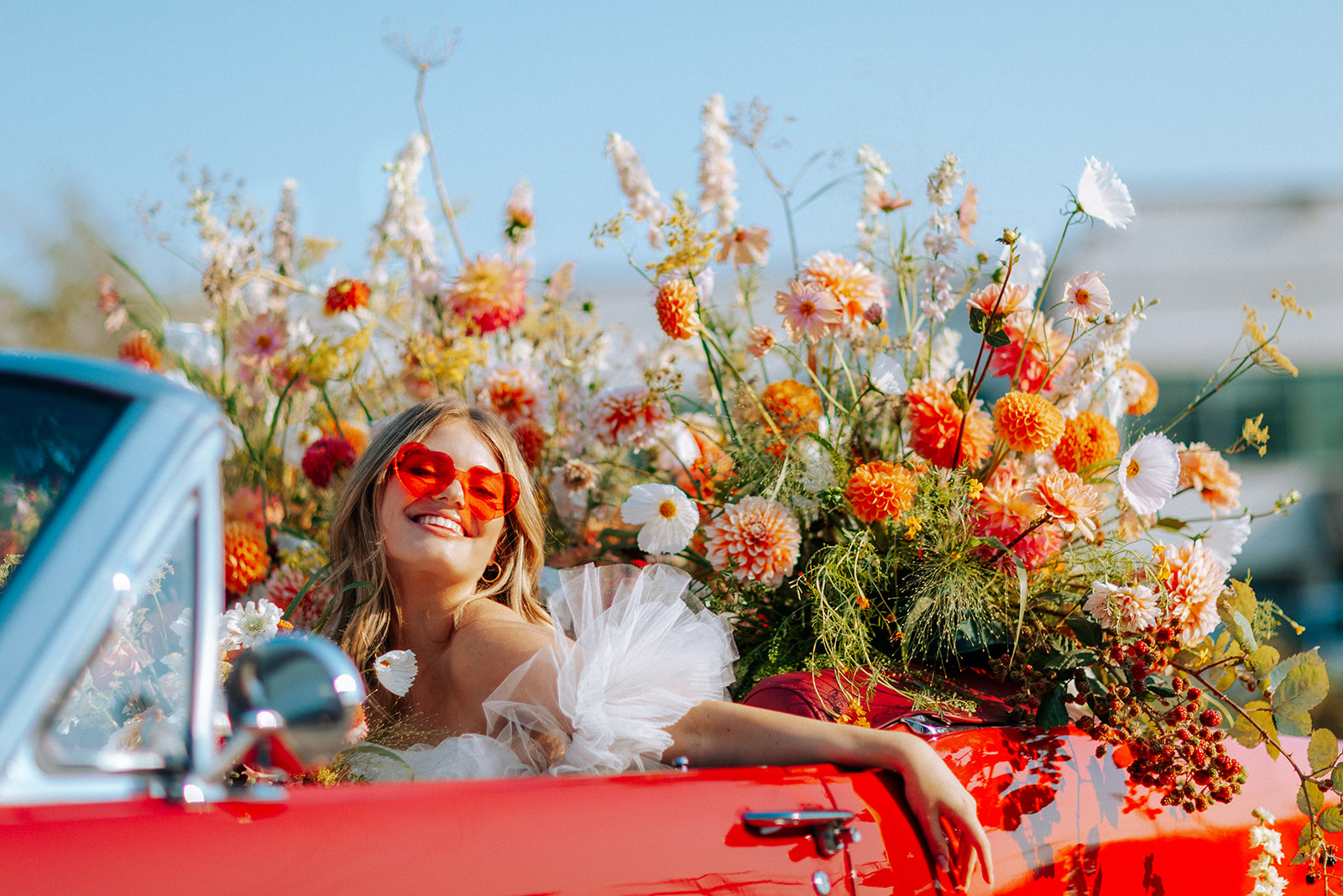 Carefree bad-ass bride wearing flirty Zephyr dress by local designer Ouma Bridal, sitting in vintage red muscle car surrounded by tangerine and white summer blooms. 