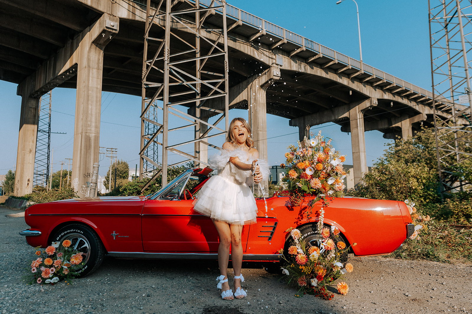 Carefree bad-ass bride wearing flirty Zephyr dress by local designer Ouma Bridal and long cathedral veil blowing in the wind, standing in front of vintage red muscle car surrounded by tangerine and white summer blooms. 
