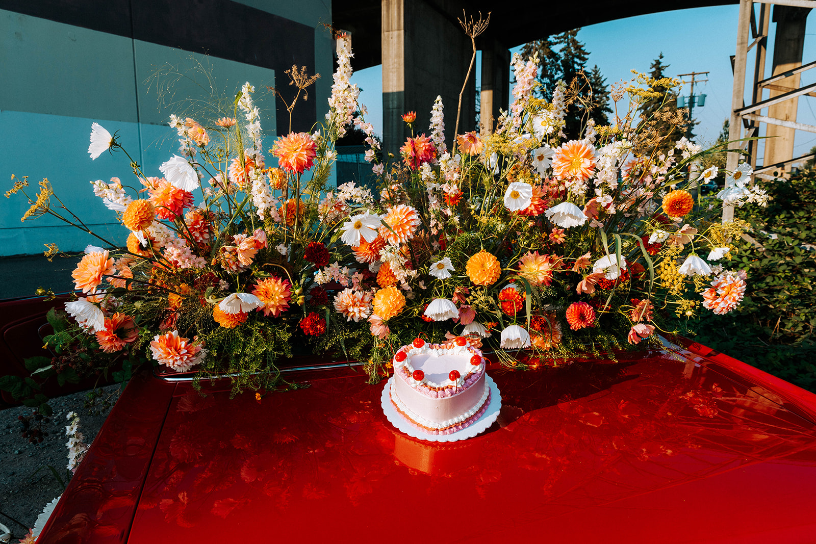 Vintage inspired pink heart cake with whipped cream and cherries, on top of vintage red muscle car. Surrounded by summer florals in white, tangerine, red, peach, and pink. 