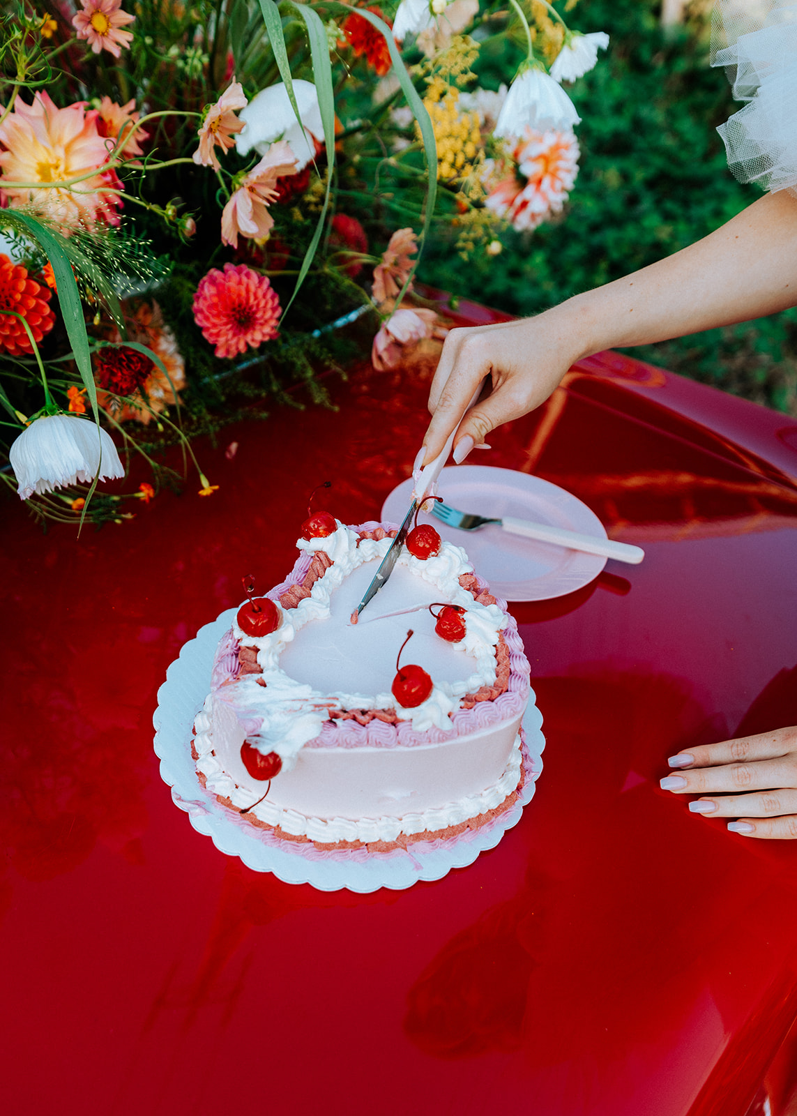 Vintage inspired pink heart cake with whipped cream and cherries, on top of vintage red muscle car. Surrounded by summer florals in white, tangerine, red, peach, and pink. 