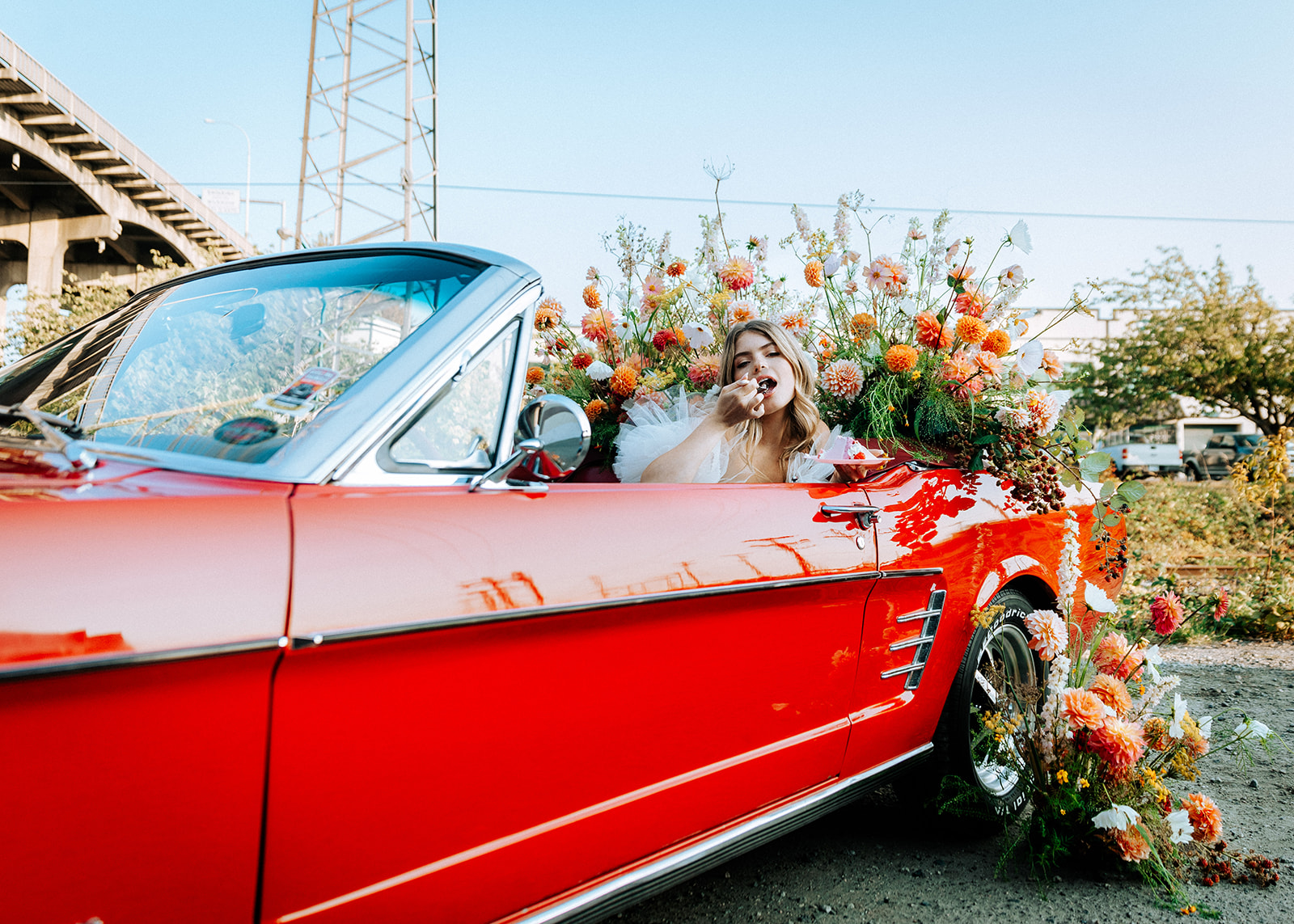 Carefree bad-ass bride wearing flirty Zephyr dress by local designer Ouma Bridal, sitting in vintage red muscle car surrounded by tangerine and white summer blooms. Poloroid image with vintage rock and roll vibes. 