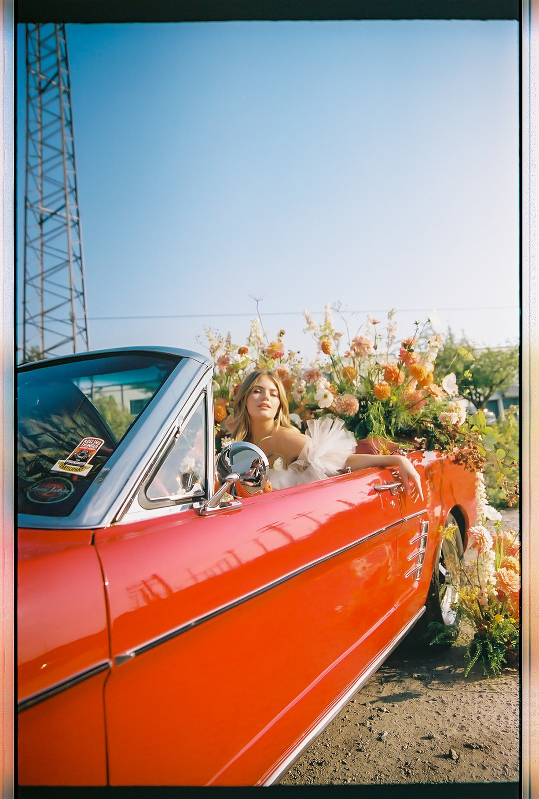 Carefree bad-ass bride wearing flirty Zephyr dress by local designer Ouma Bridal, sitting in vintage red muscle car surrounded by tangerine and white summer blooms. Poloroid image with vintage rock and roll vibes. 