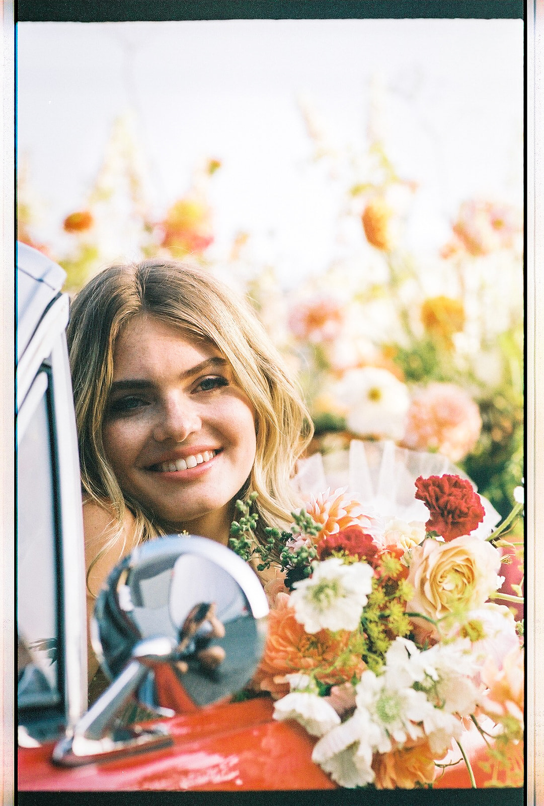 Carefree bad-ass bride wearing flirty Zephyr dress by local designer Ouma Bridal, sitting in vintage red muscle car surrounded by tangerine and white summer blooms. Poloroid image with vintage rock and roll vibes. 