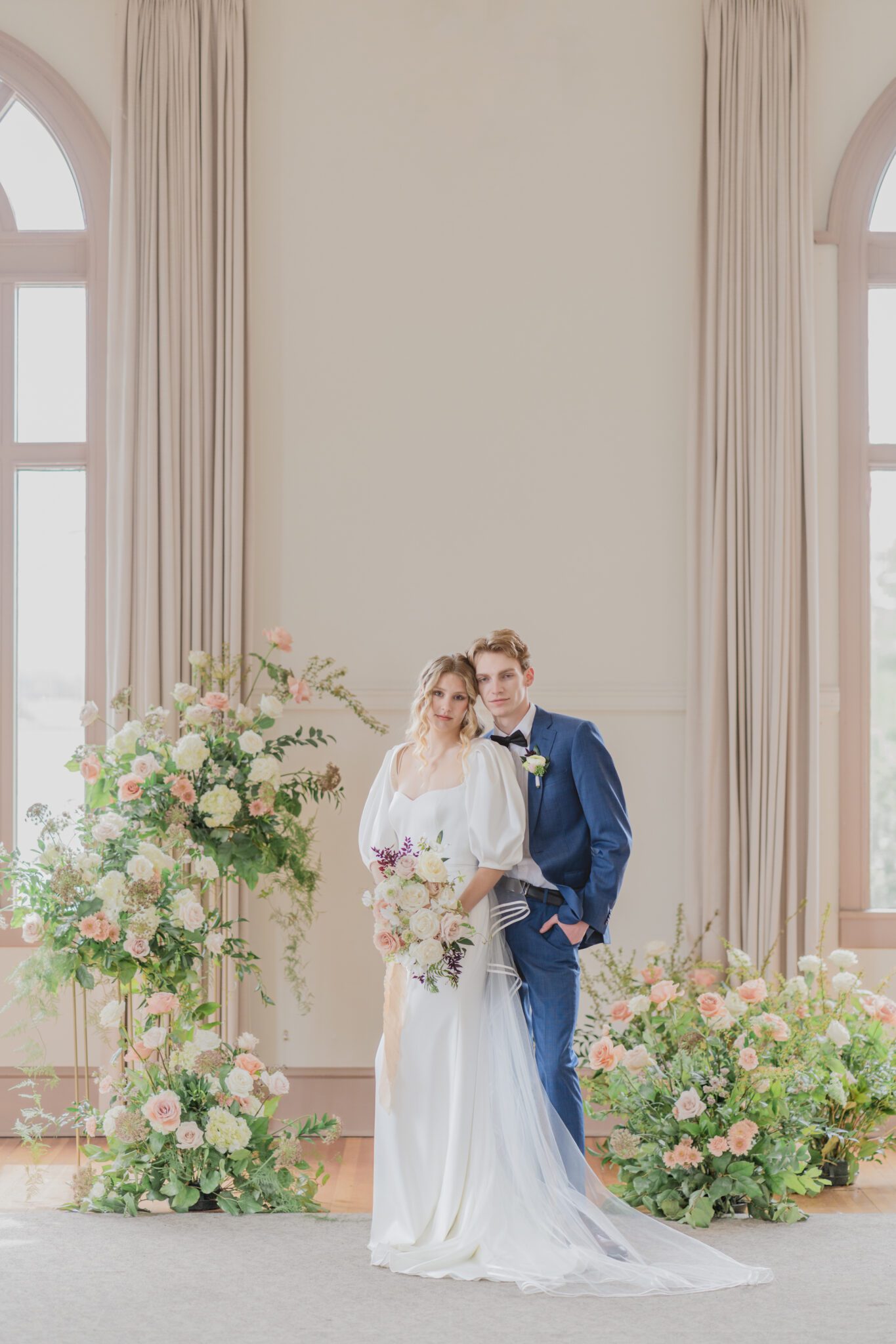 Bride and groom during timeless elegant wedding at Chilliwack Museum in front of ceremony arch with cascading floral arrangements in white and peach with touches of gold. Bride wearing an elegant crepe wedding dress with a removable bolero and sweetheart neckline. 