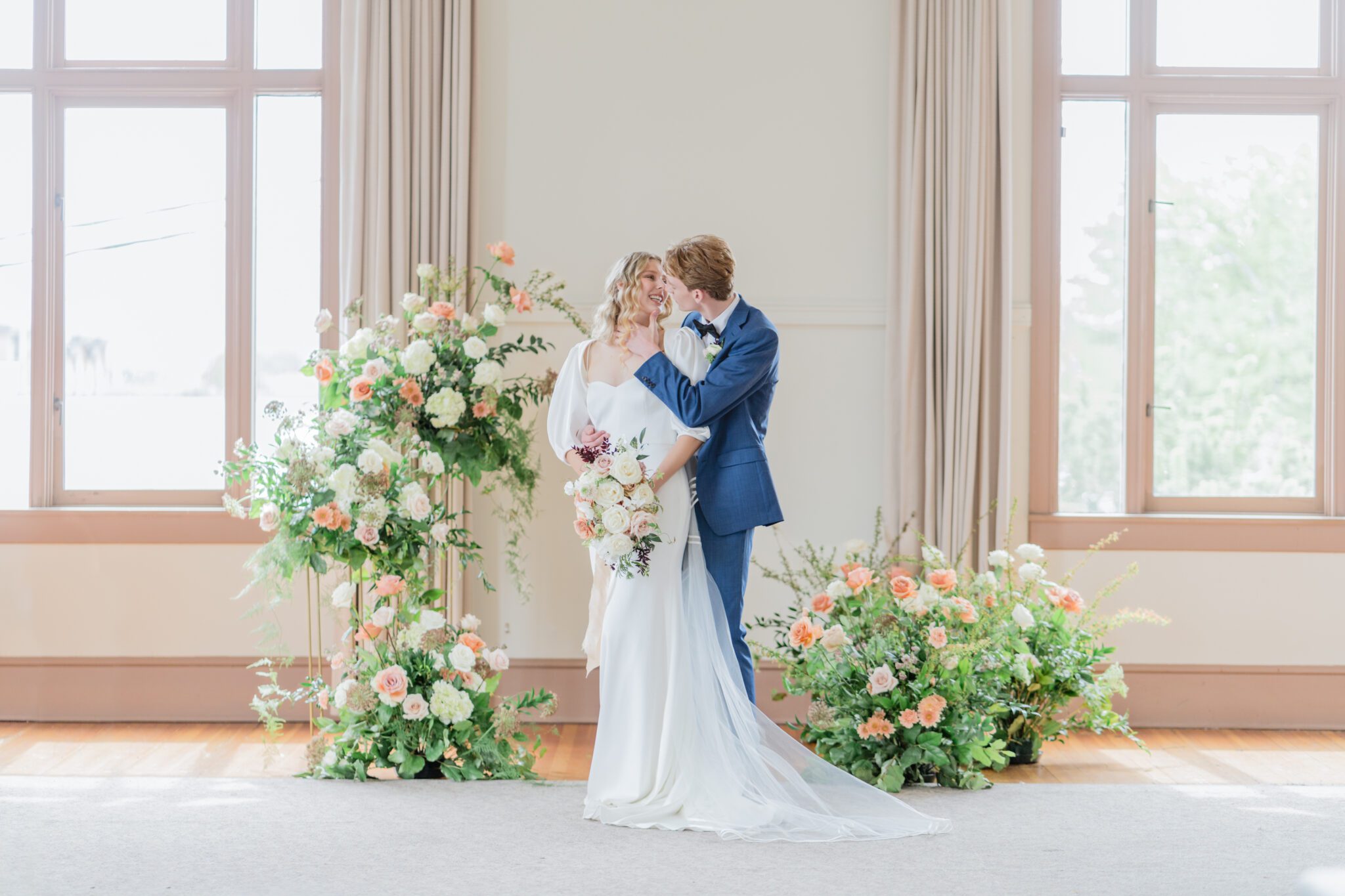 Bride and groom during timeless elegant wedding at Chilliwack Museum in front of ceremony arch with cascading floral arrangements in white and peach with touches of gold. Bride wearing an elegant crepe wedding dress with a removable bolero and sweetheart neckline. 