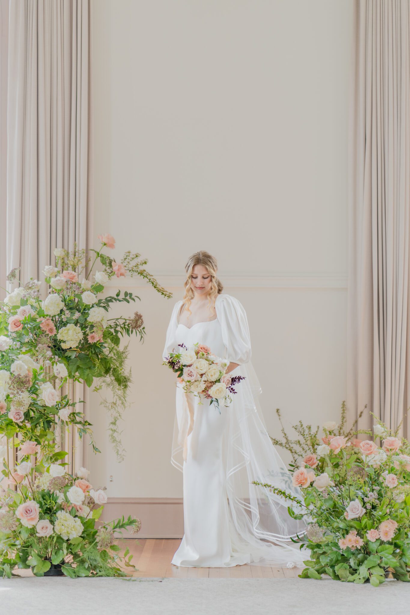 Timeless elegant wedding at Chilliwack Museum in front of ceremony arch with cascading floral arrangements in white and peach with touches of gold. Bride wearing an elegant crepe wedding dress with a removable bolero and sweetheart neckline. 