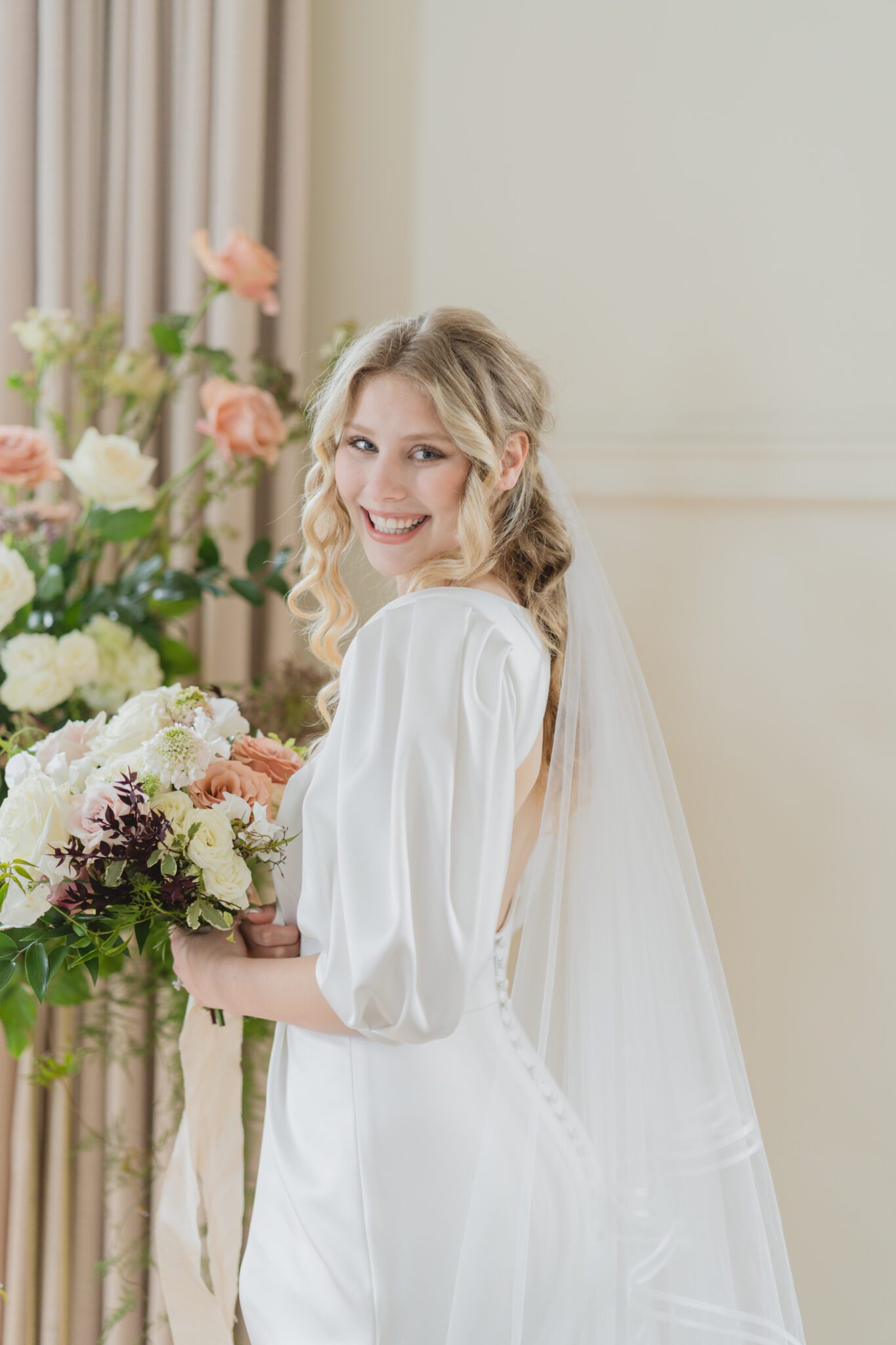 Timeless elegant wedding at Chilliwack Museum in front of ceremony arch with cascading floral arrangements in white and peach with touches of gold. Bride wearing an elegant crepe wedding dress with a removable bolero and sweetheart neckline. 