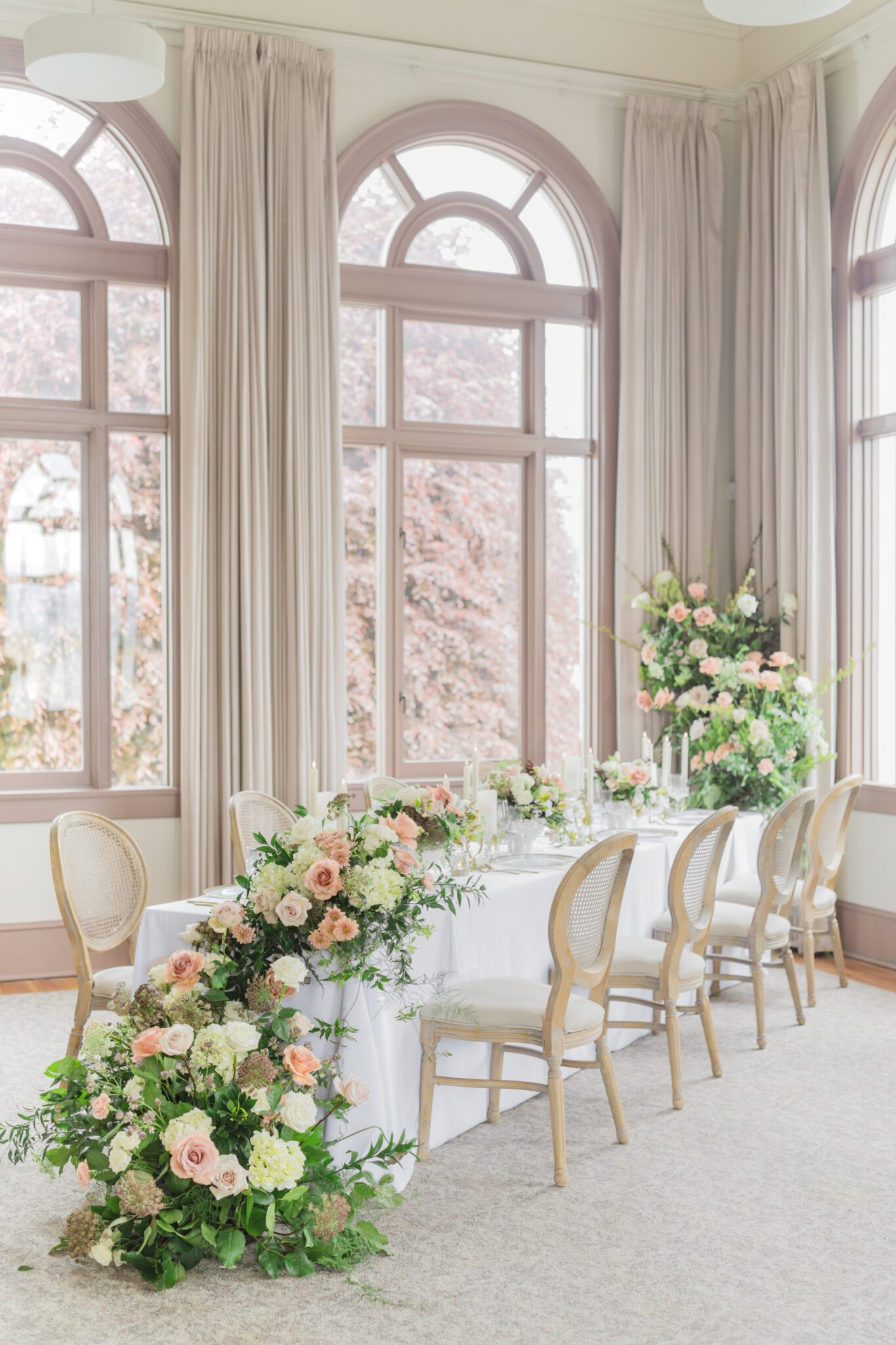 Wedding reception table at Chilliwack Museum, adorned with luxurious gold cutlery, unique candleholders, and vintage-inspired glasses, complemented the cane back chairs. Cascading floral centrepiece and arrangements in white and peach.