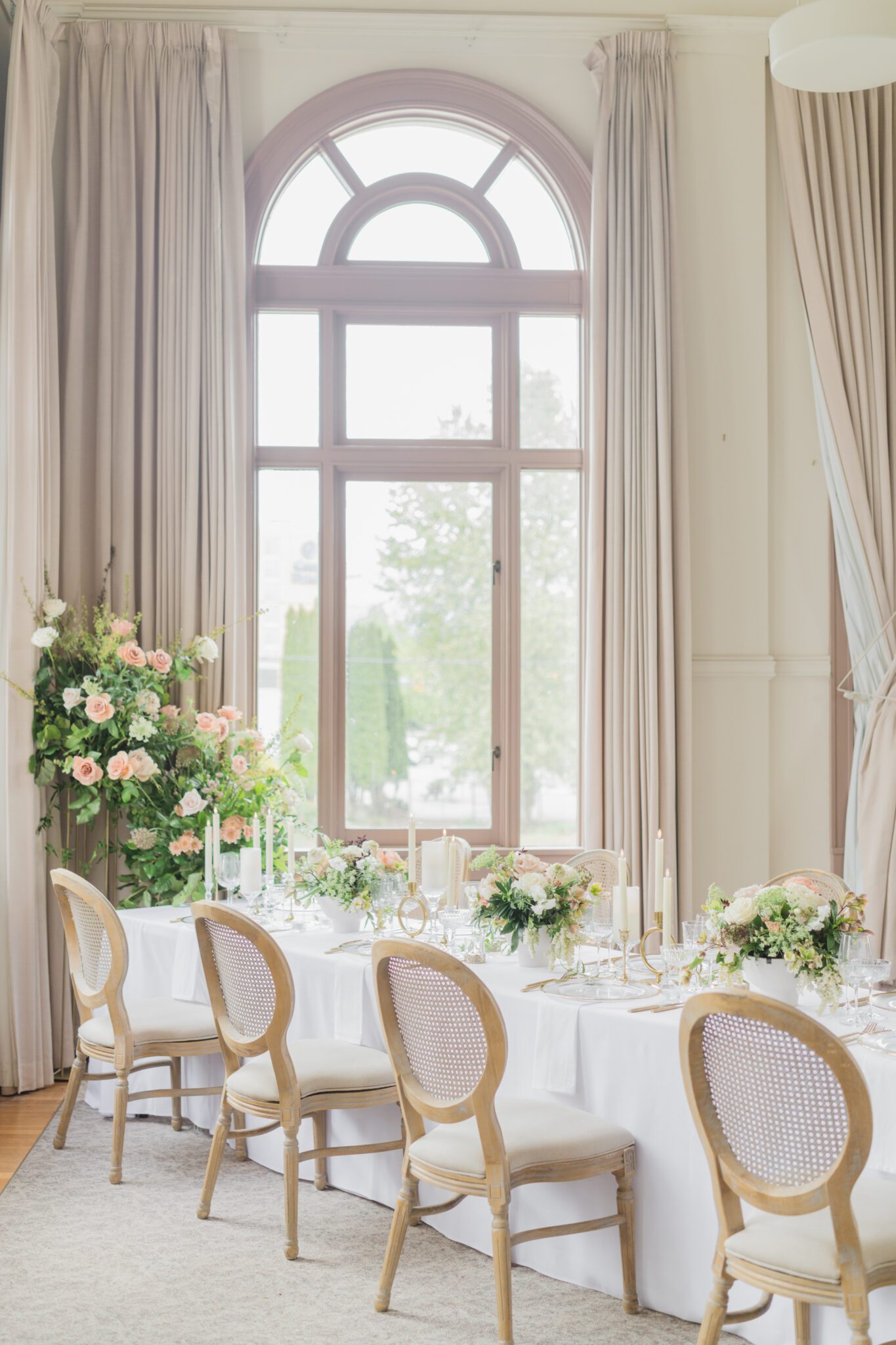 Wedding reception table at Chilliwack Museum, adorned with luxurious gold cutlery, unique candleholders, and vintage-inspired glasses, complemented the cane back chairs. Cascading floral centrepiece and arrangements in white and peach.