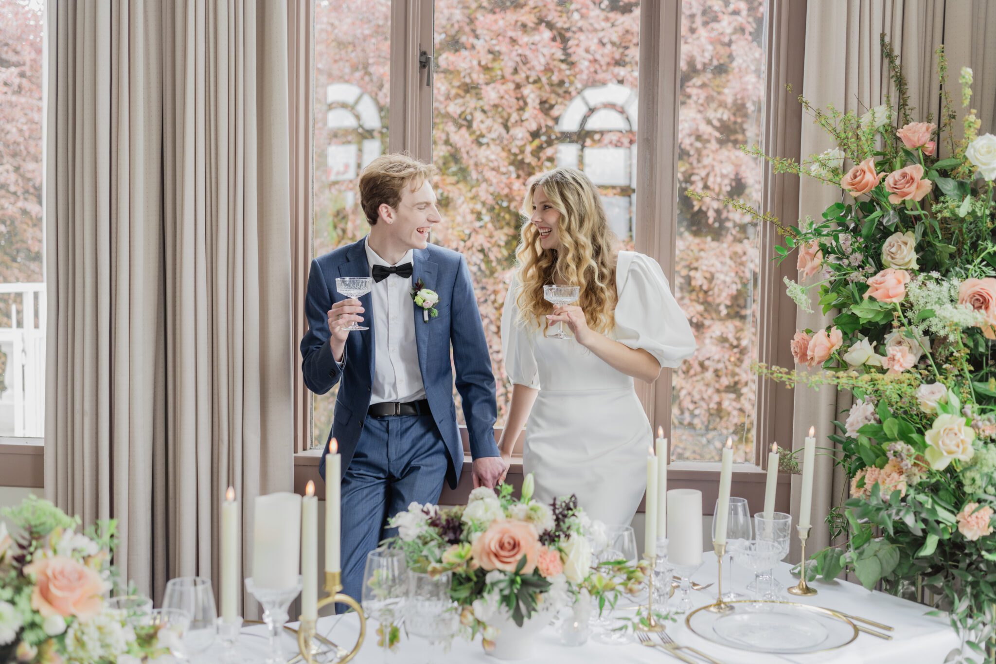 Bride and groom toasting during wedding reception at Chilliwack Museum. Table adorned with luxurious gold cutlery, unique candleholders, and vintage-inspired glasses, complemented the cane back chairs. Cascading floral centrepiece and arrangements in white and peach.