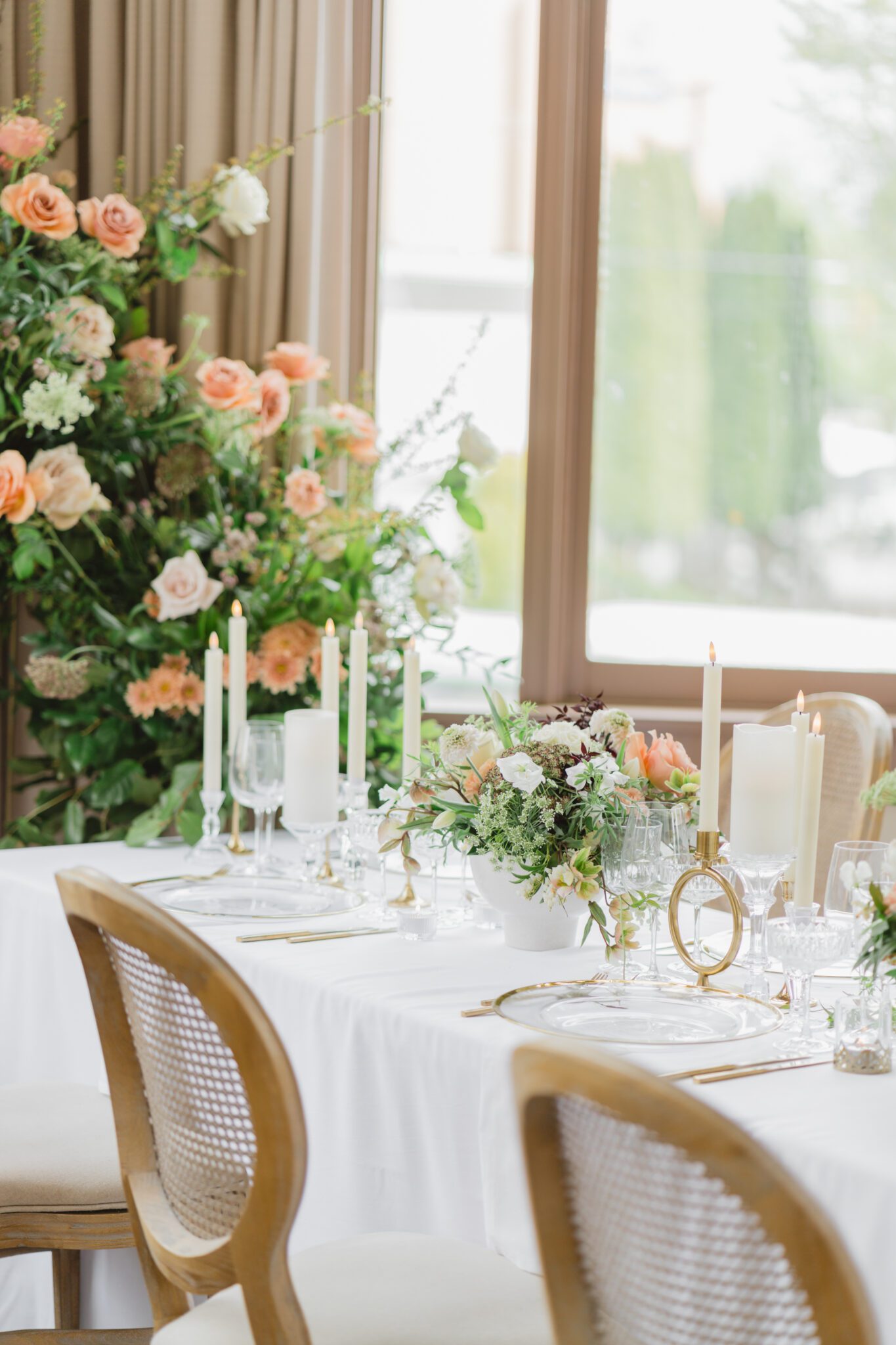 Wedding reception table at Chilliwack Museum, adorned with luxurious gold cutlery, unique candleholders, and vintage-inspired glasses, complemented the cane back chairs. Cascading floral centrepiece and arrangements in white and peach.