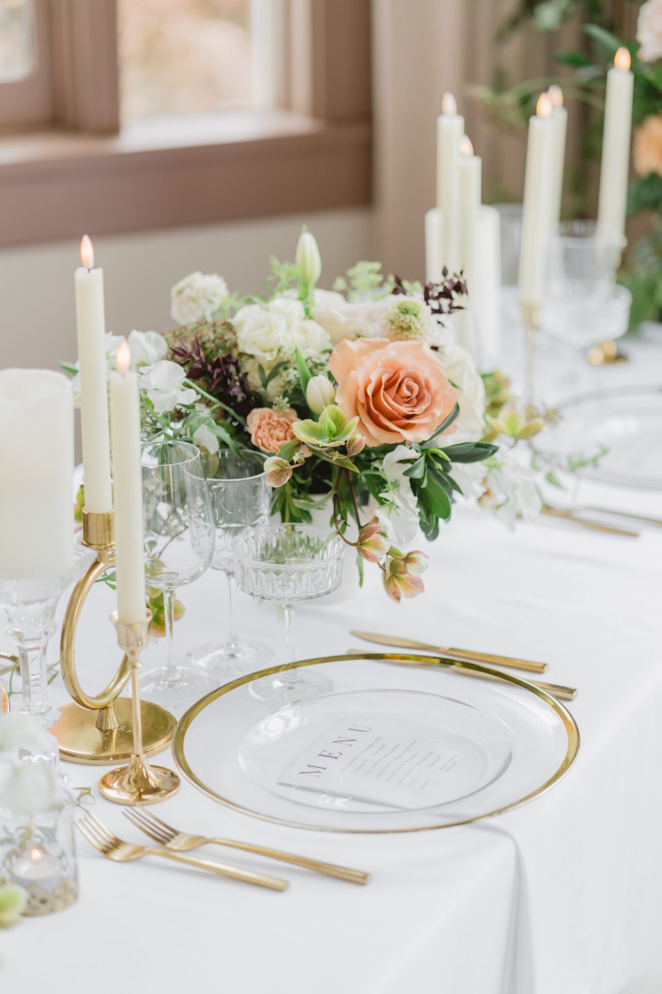 Wedding reception table at Chilliwack Museum, adorned with luxurious gold cutlery, unique candleholders, and vintage-inspired glasses, complemented the cane back chairs. Cascading floral centrepiece and arrangements in white and peach.