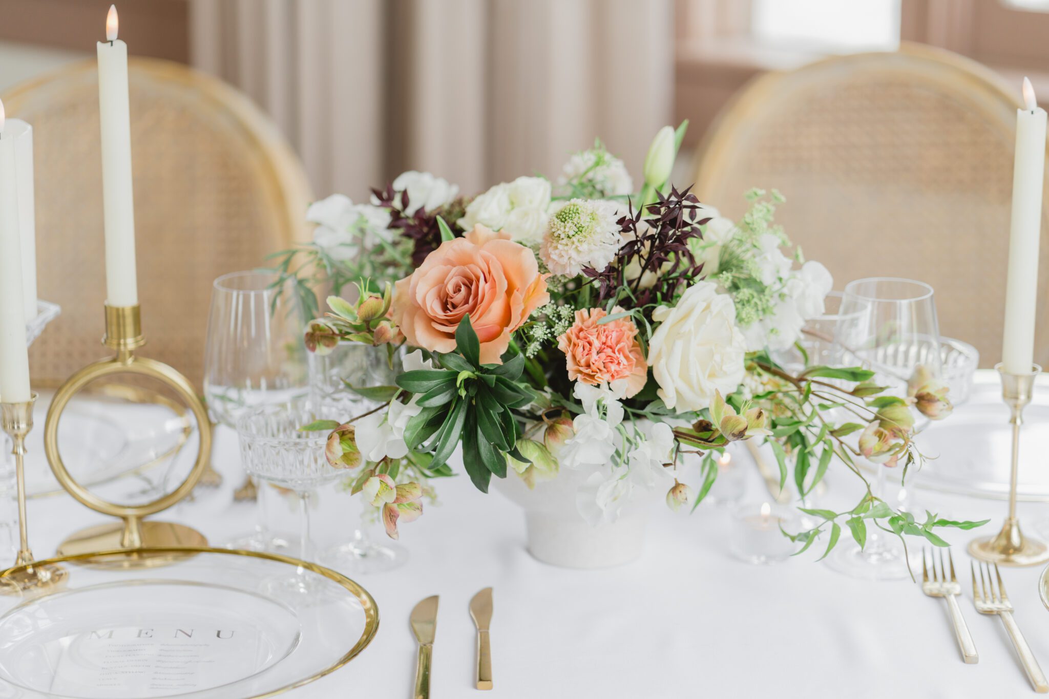 Wedding reception table at Chilliwack Museum, adorned with luxurious gold cutlery, unique candleholders, and vintage-inspired glasses, complemented the cane back chairs. Cascading floral centrepiece and arrangements in white and peach.