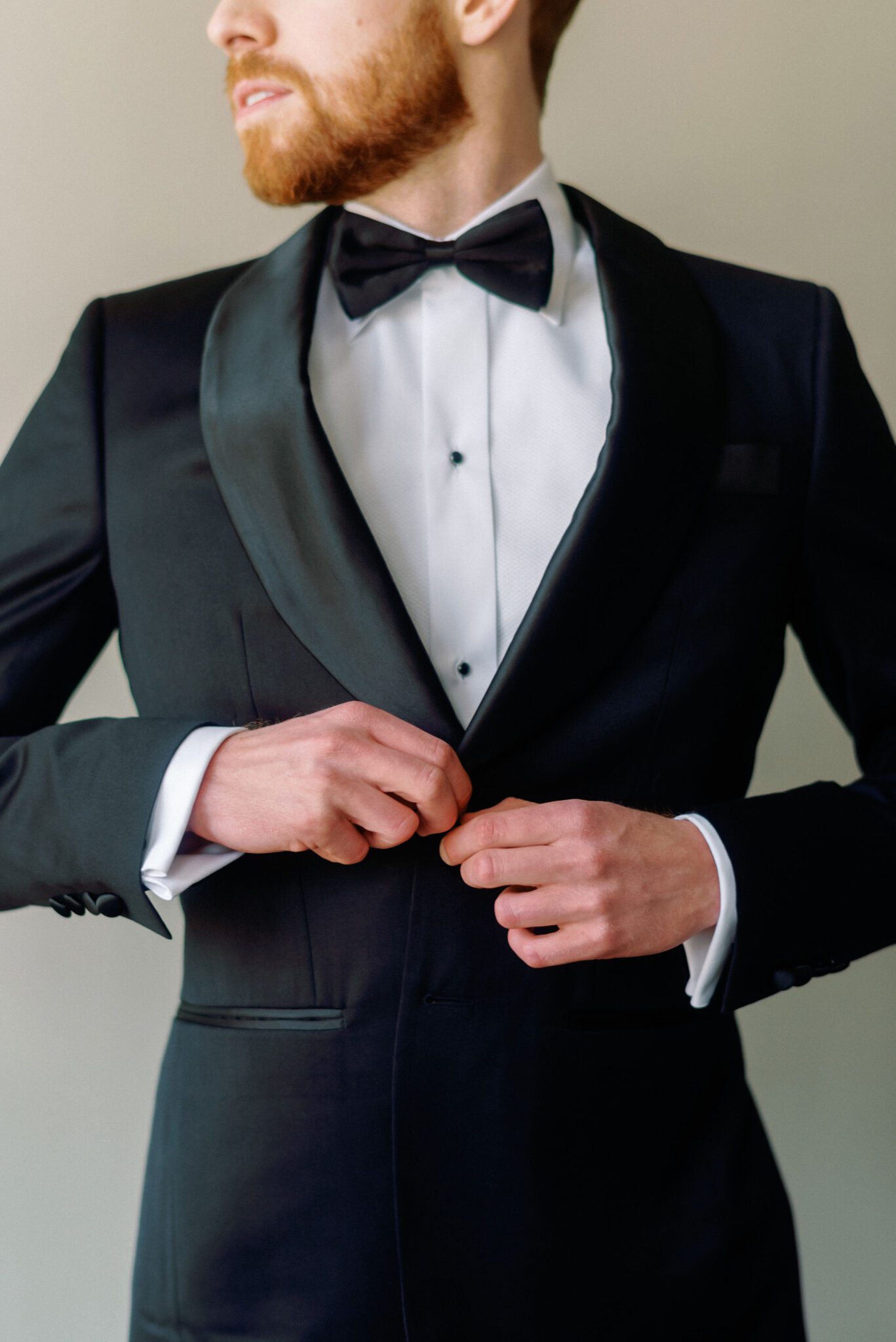 Groom getting ready in classic black suit with satin lapel, crisp white shirt with black buttons, and black bow tie creating a suave look for the modern groom. 