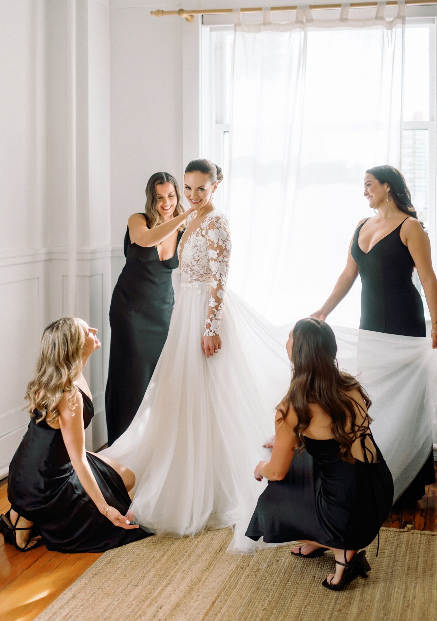 Bride surrounded by bridesmaids in sleek black satin dresses, helping her with the final touches on her flowing white gown. The bride's lace wedding dress with sheer sleeves and intricate floral details stands out against the minimalistic backdrop.