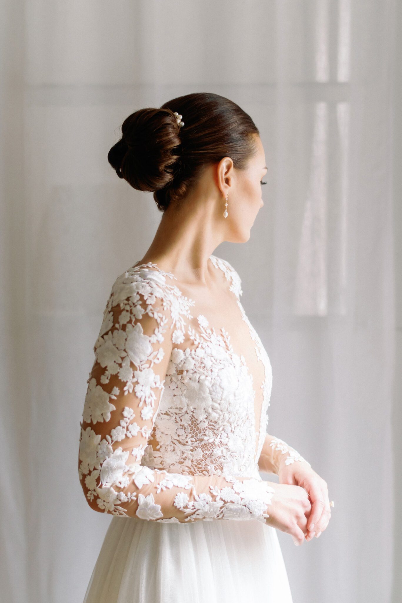 Elegant bride in a long-sleeve lace wedding gown with intricate floral embroidery. The delicate sheer sleeves and deep V-neckline create a romantic, sophisticated look. Her hair is styled in a sleek low bun, adorned with pearls, complementing the timeless elegance of the dress. 