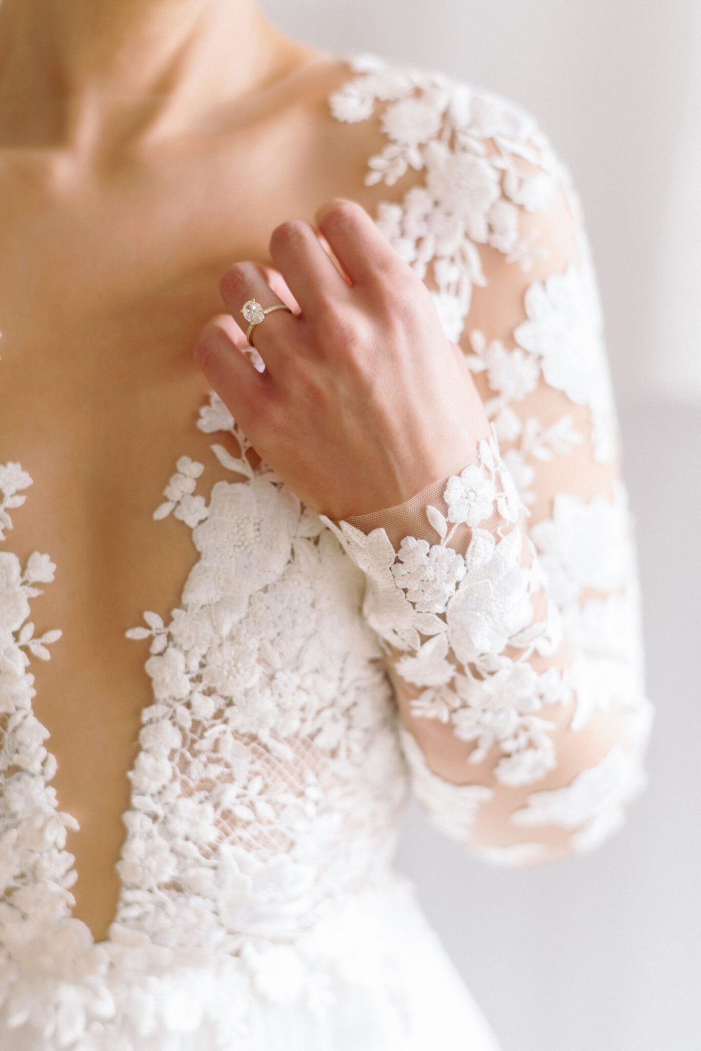 Elegant bride in a long-sleeve lace wedding gown with intricate floral embroidery. The delicate sheer sleeves and deep V-neckline create a romantic, sophisticated look. Her hair is styled in a sleek low bun, adorned with pearls, complementing the timeless elegance of the dress. 