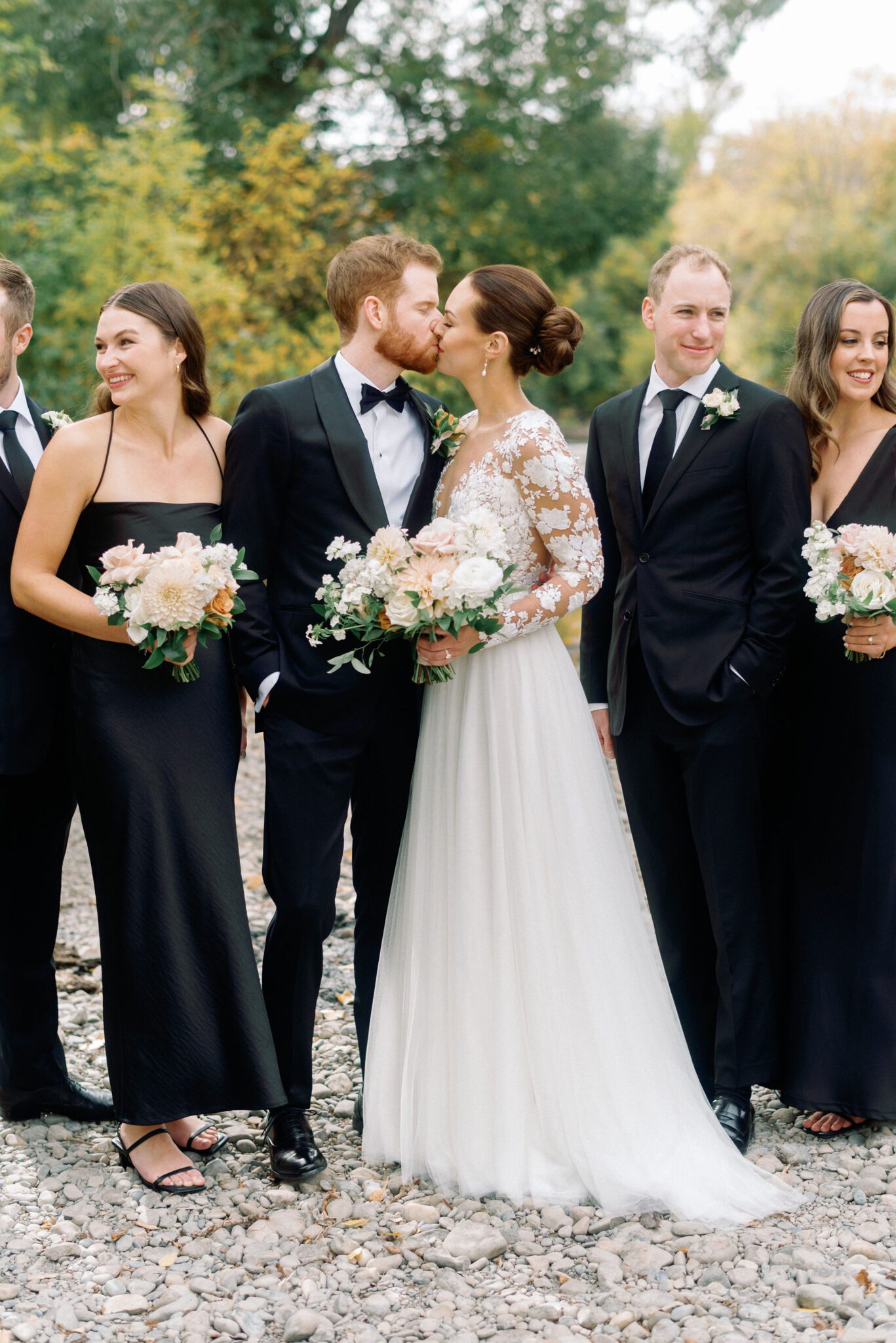 Bride and groom kiss during a romantic wedding portrait session surrounded by wedding party. 