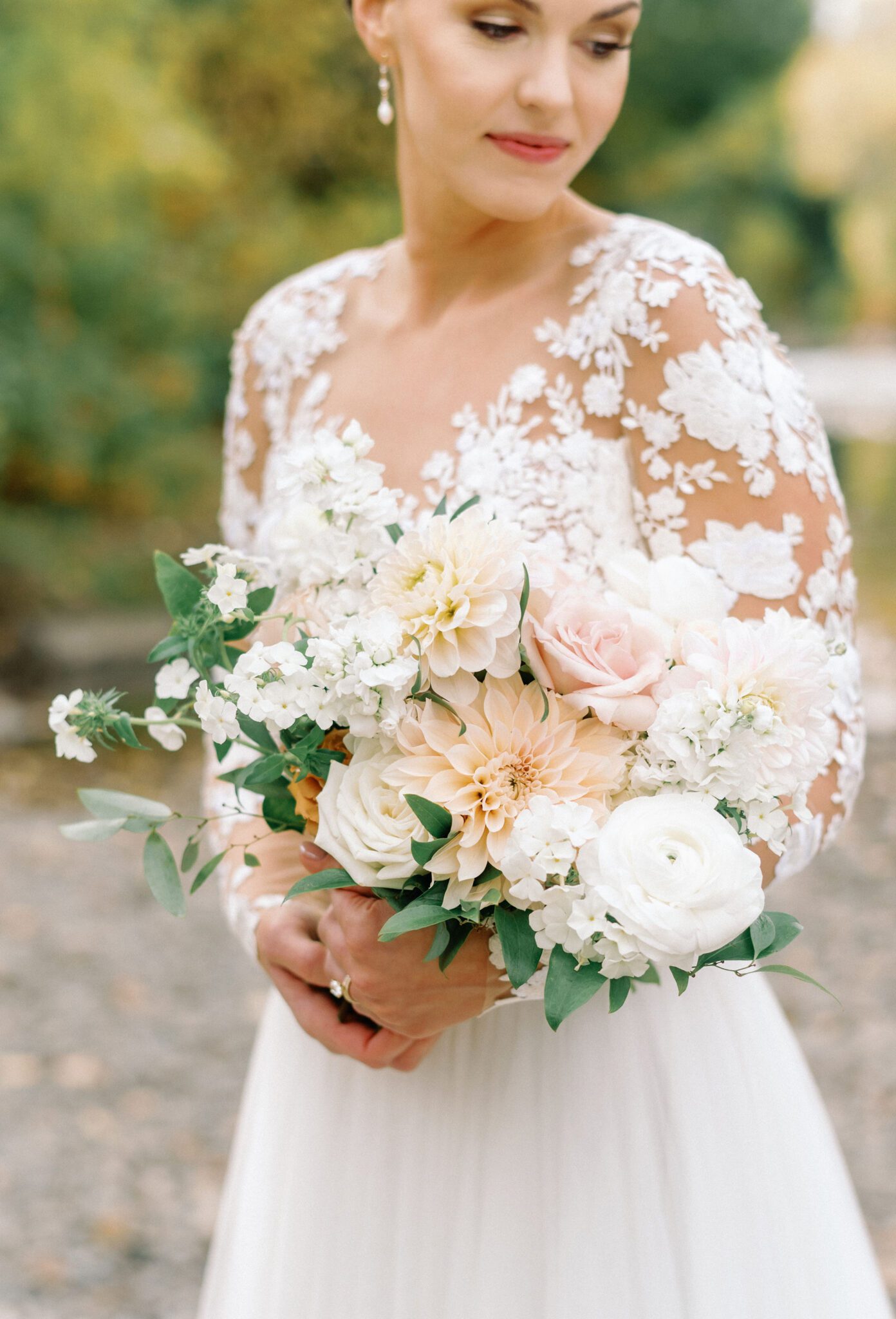 Elegant bride in a long-sleeve lace wedding gown with intricate floral embroidery. The delicate sheer sleeves and deep V-neckline create a romantic, sophisticated look. Her hair is styled in a sleek low bun, adorned with pearls, complementing the timeless elegance of the dress. 