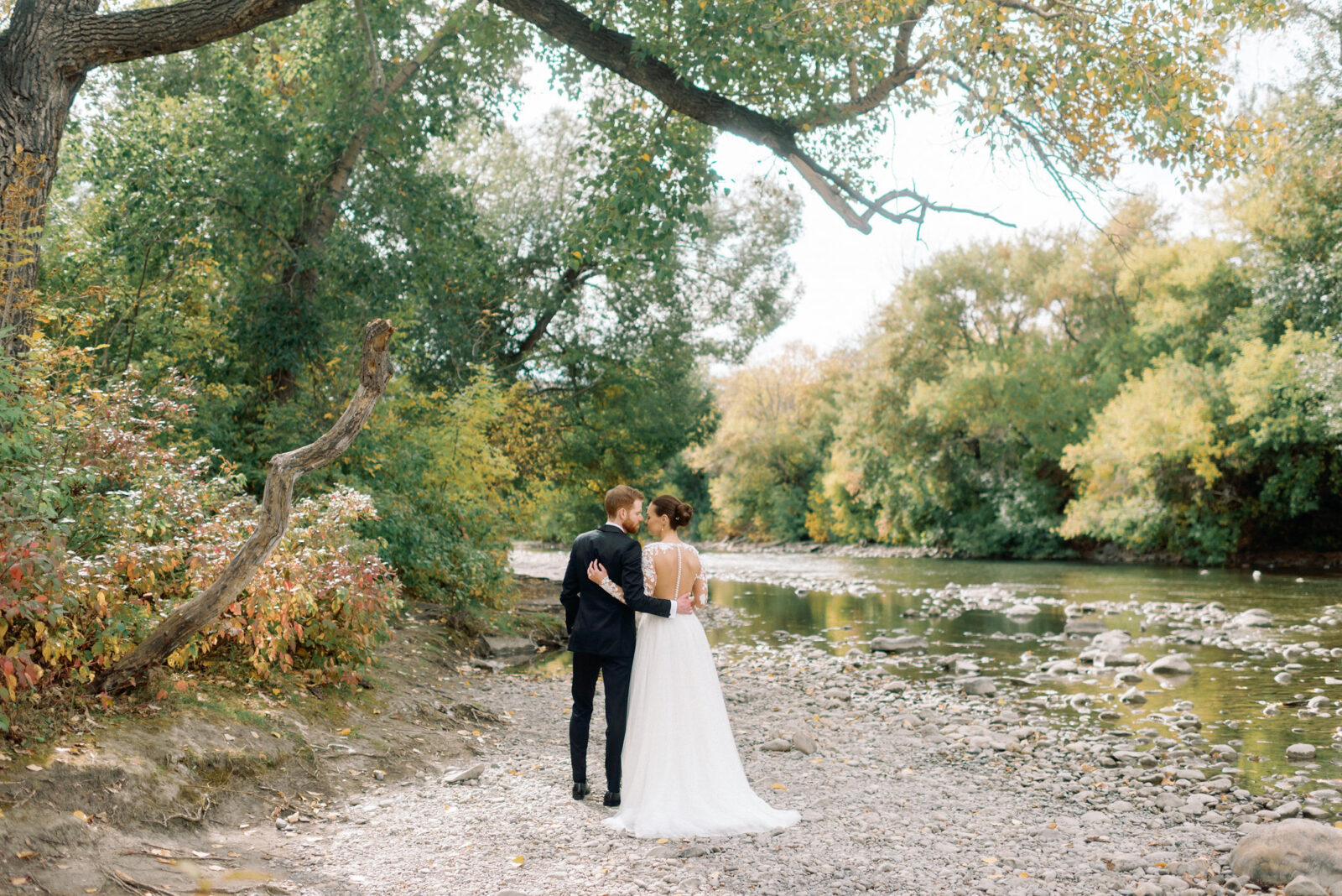 Bride and groom embrace during a romantic wedding portrait session by the river surrounded by trees.