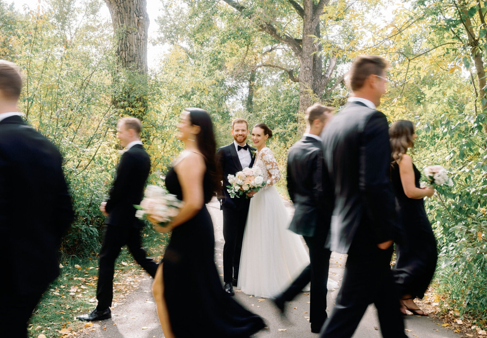 Bride and groom embrace during a fun wedding portrait session, bridesmaids. and groomsmen walking in foreground. 