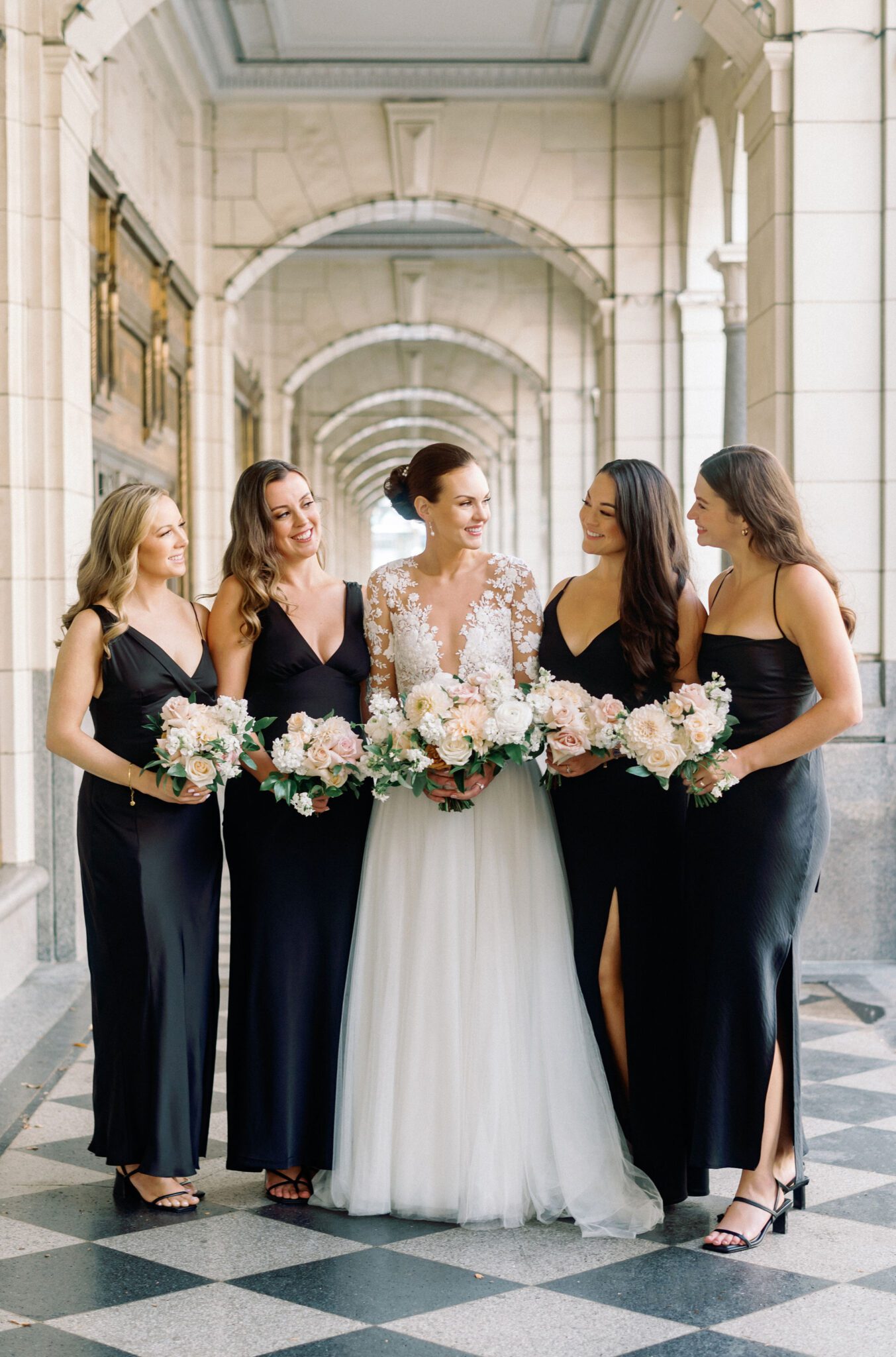 Bride surrounded by bridesmaids in sleek black satin dresses. The bride's lace wedding dress with sheer sleeves and intricate floral details stands out against the architectural backdrop. 