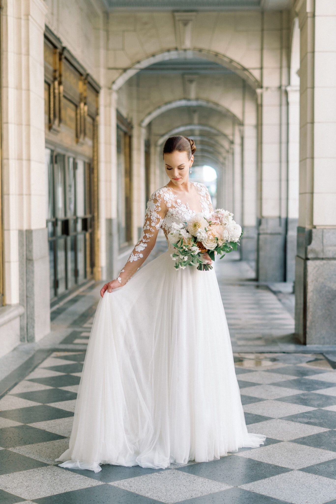 Elegant bride in a long-sleeve lace wedding gown with intricate floral embroidery. The delicate sheer sleeves and deep V-neckline create a romantic, sophisticated look. Her hair is styled in a sleek low bun, adorned with pearls, complementing the timeless elegance of the dress. 