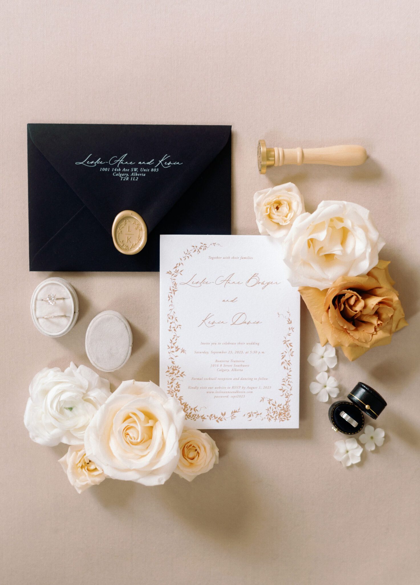 Elegant wedding invitation set with black envelope, gold wax seal, and white floral accents. The invitation features delicate gold leaf details and a formal script, creating a luxurious and timeless design. A diamond engagement ring in a white velvet box and soft white roses complement the stationery, emphasizing a classic, romantic aesthetic.