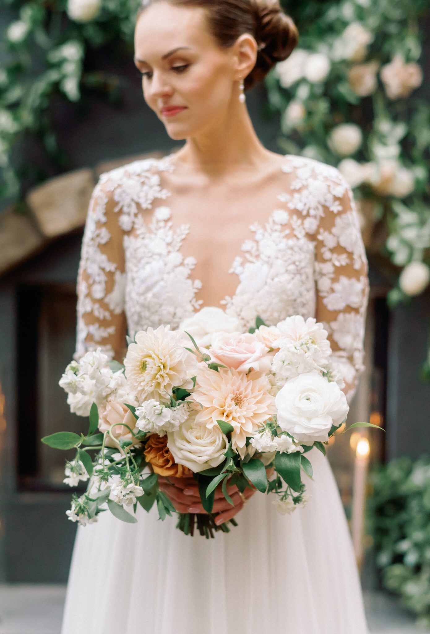 Elegant bride in a long-sleeve lace wedding gown with intricate floral embroidery. The delicate sheer sleeves and deep V-neckline create a romantic, sophisticated look. Her hair is styled in a sleek low bun, adorned with pearls, complementing the timeless elegance of the dress. 