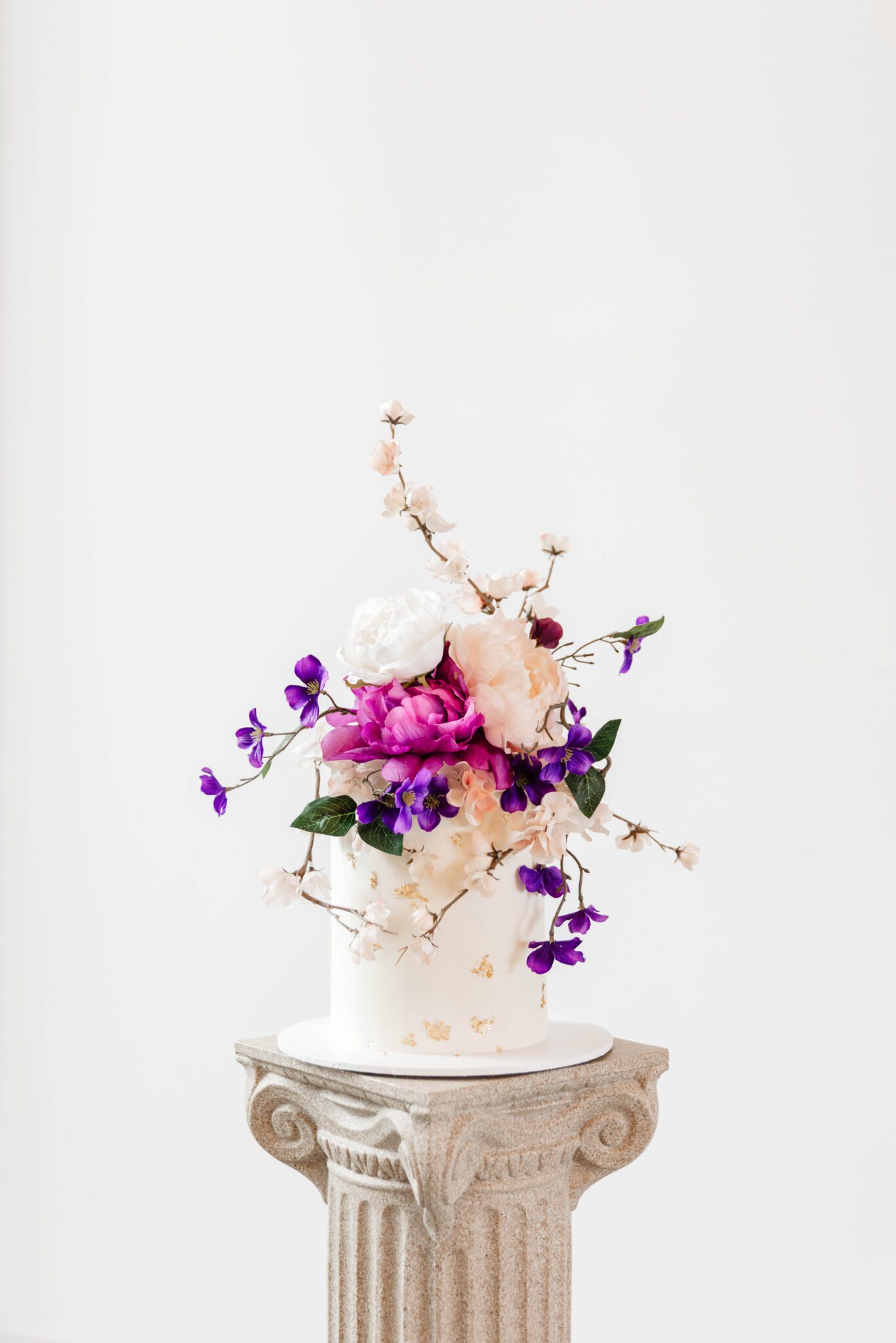 Elegant one-tiered wedding cake topped with purple, white, and pink peonies, cherry blossoms, and delicate purple garden flowers. 