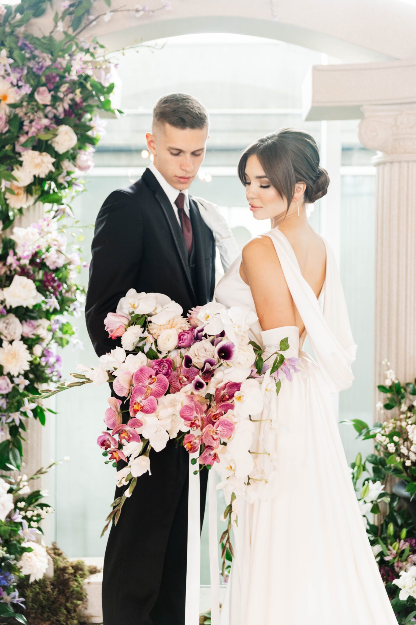 Couple during purple garden wedding ceremony at historic venue The Pendennis in Edmonton, Alberta. Lush purple garden-inspired florals. Architectural column archway, with cascading floral installation. 