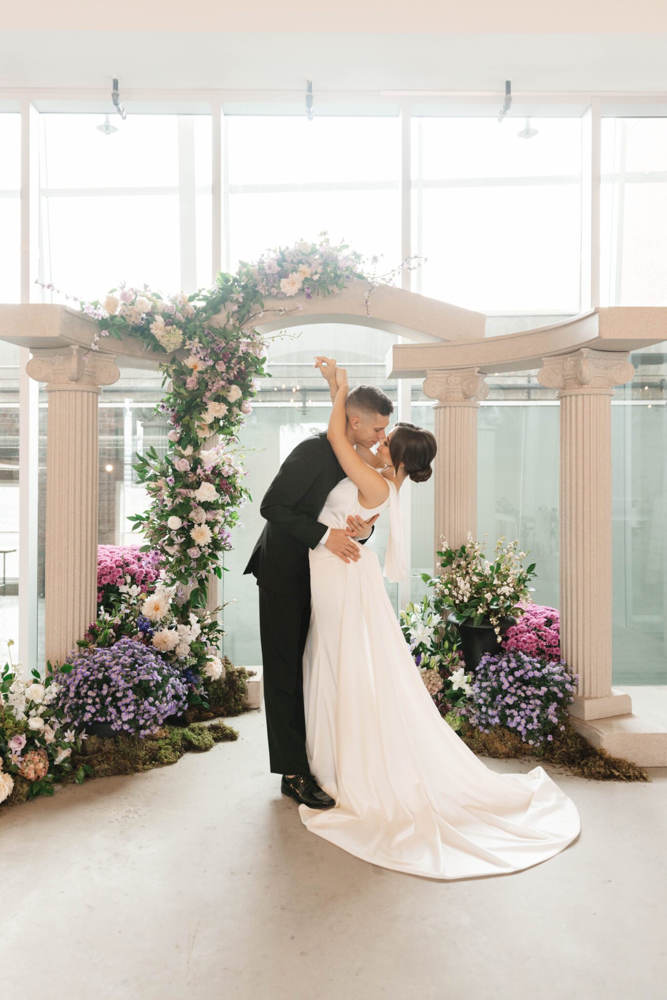 Couple kissing during purple Garden Wedding at historic venue The Pendennis in Edmonton, Alberta. Lush purple garden-inspired florals. Architectural column archway, with cascading floral installation. 