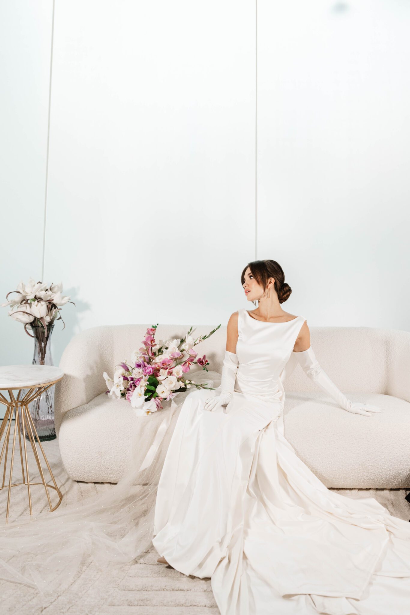 Elegant bride sitting on sofa at The Pendennis in Edmonton, Alberta. Elegant bridal bouquet of white orchids, gladiolas, and purple garden flowers. 