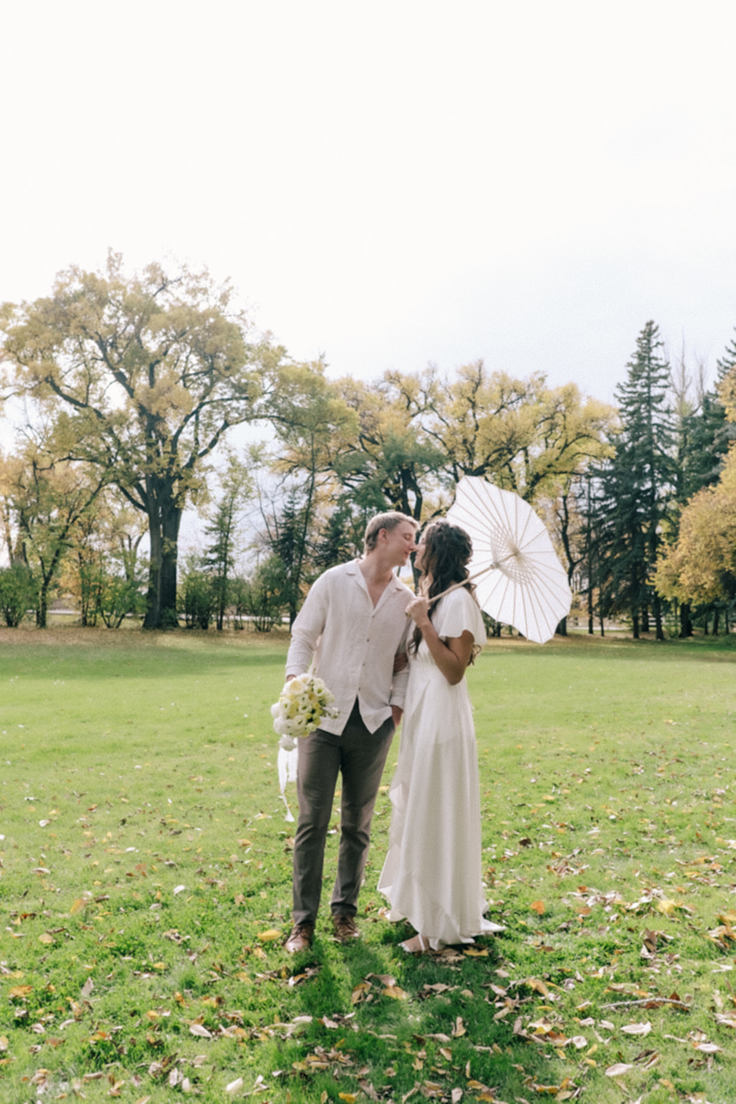 Bride and groom share a romantic moment under a canopy of trees during a summer wedding. Featuring outdoor portraits with white paper parasol and pale yellow bouquet.