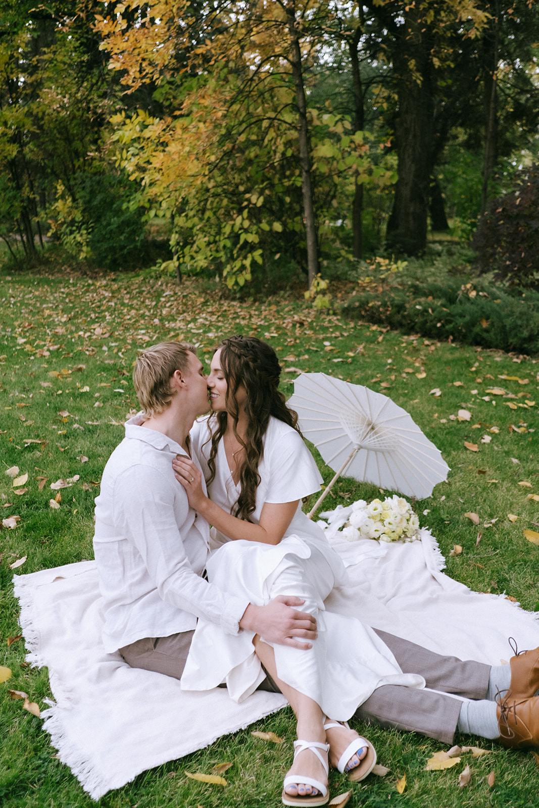 Bride and groom share a romantic moment under a canopy of trees during a summer wedding. Featuring outdoor portraits with white paper parasol and pale yellow bouquet.