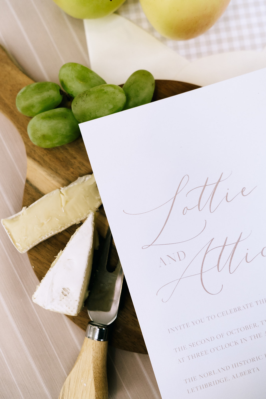 Close-up of wedding invitation flatlay surrounded by fruit and cheese on a wood cutting board, embracing the cottage core aesthetic.