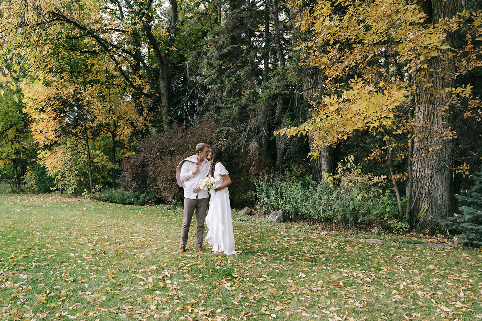 Bride and groom share a romantic moment under a canopy of trees at a casual backyard wedding with a cottage core theme. Bride wearing simple sink gown created by By Catalfo, holding a bouquet of pale yellow flowers. 
