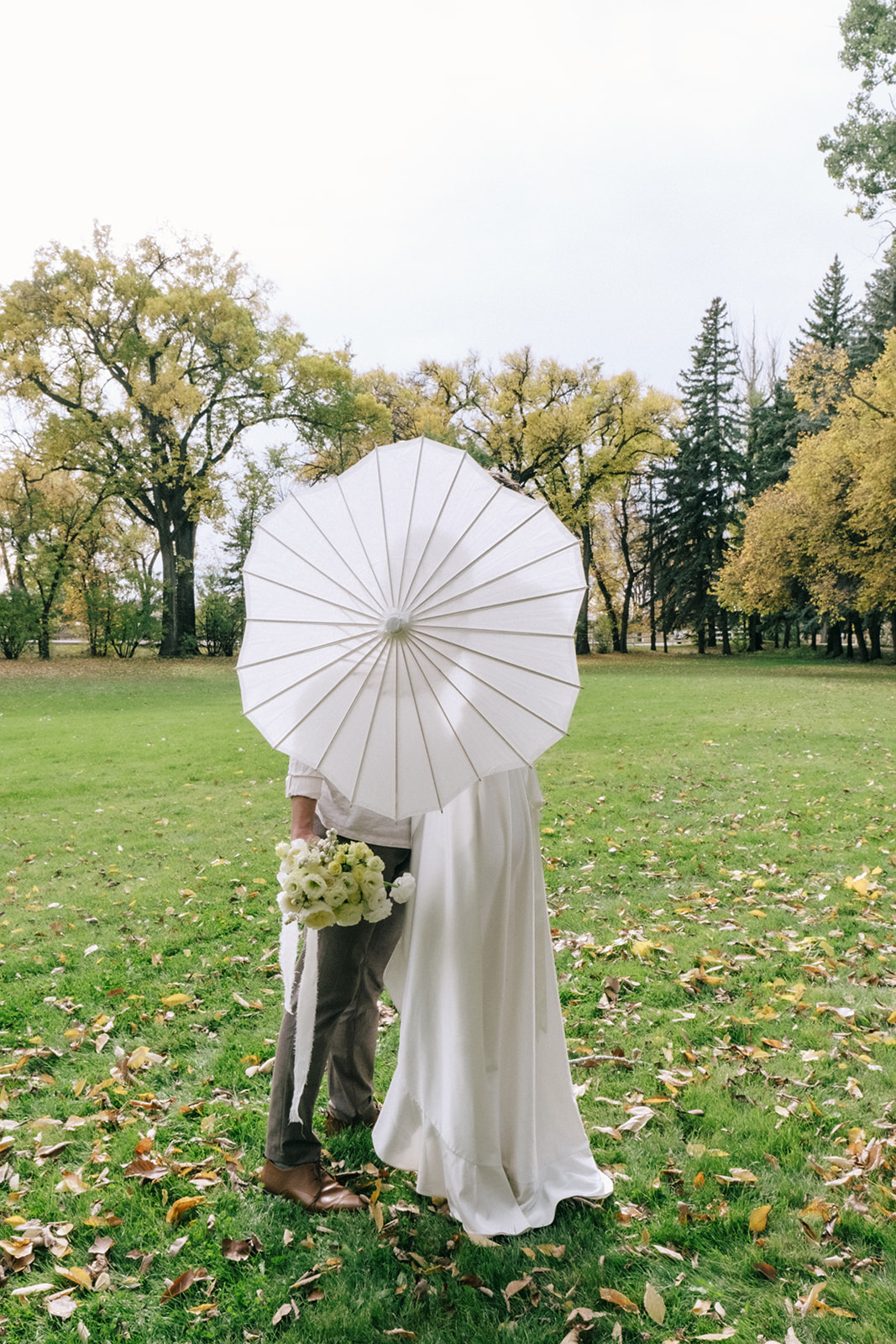 Bride and groom share a romantic moment under a canopy of trees during a summer wedding. Featuring outdoor portraits with white paper parasol and pale yellow bouquet.