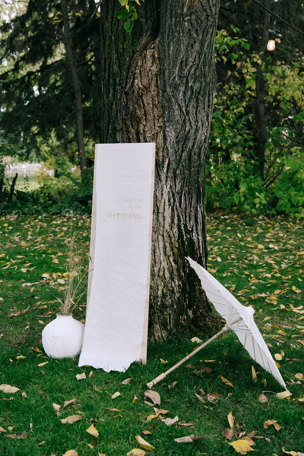 Cottage core wedding ceremony welcome sign with wood and white linen textures, delicate wildflowers and vintage-inspired white wedding parasol. 