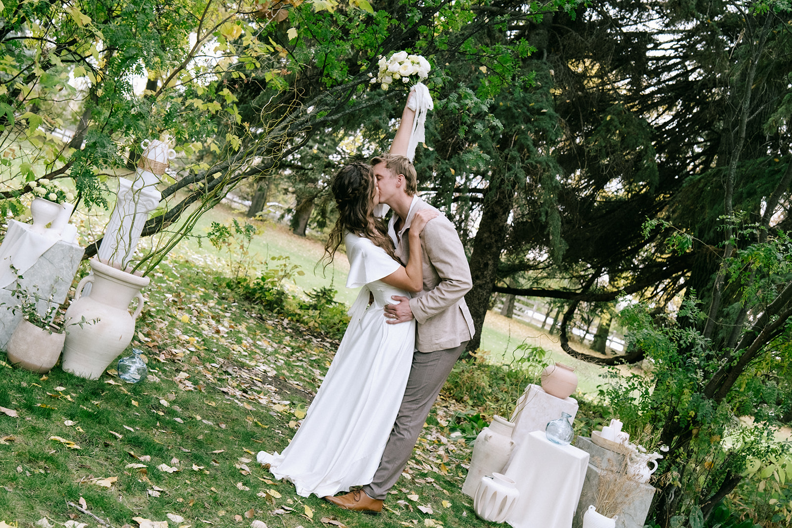 Bride and groom share their first kiss at cottage core inspired wedding ceremony at The Norland Historic Estate in Southern Alberta. Pedestal stand ceremony arch design layered with flowy fabric, organic neutral vases with florals and grasses, and candles.