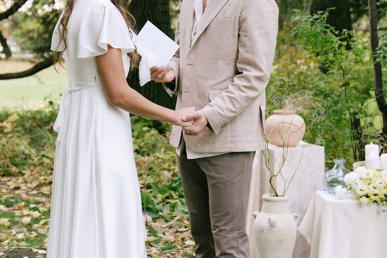 Bride and groom share their vows at cottage core inspired wedding ceremony at The Norland Historic Estate in Southern Alberta. Pedestal stand ceremony arch design layered with flowy fabric, organic neutral vases with florals, and candles. 