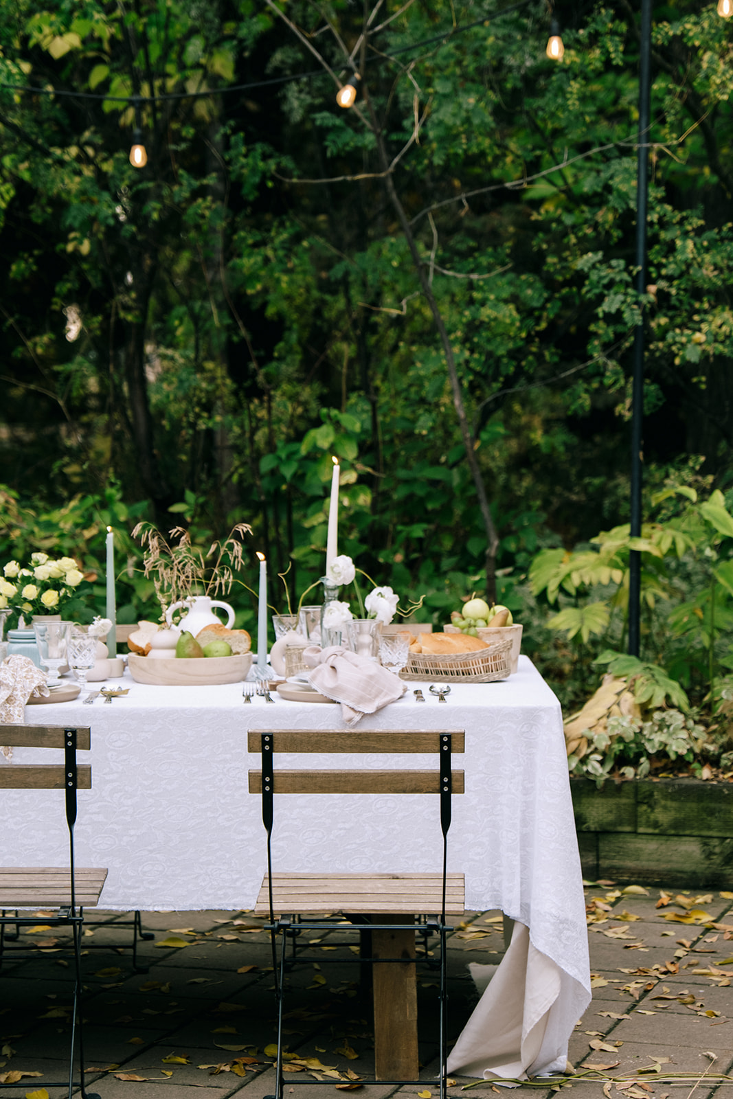 Cottage core wedding table with a subtle sheer patterned tablecloth, mist-colored taper candles, and organic floral centerpieces. Family-style wedding table setting with grapes, bread, and cheese, surrounded by earthy grasses and pale yellow blooms.