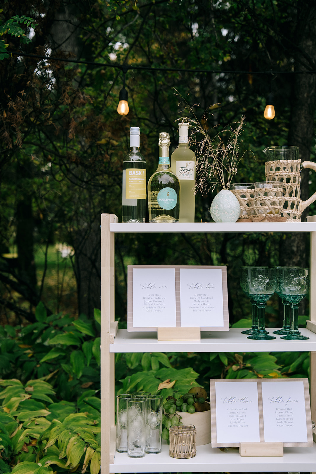 Rustic white and wood shelving display with wine bottles and vintage colored glassware for a cottage core wedding. Seating chart and drink display with mix and match glassware, perfect for a whimsical backyard wedding.