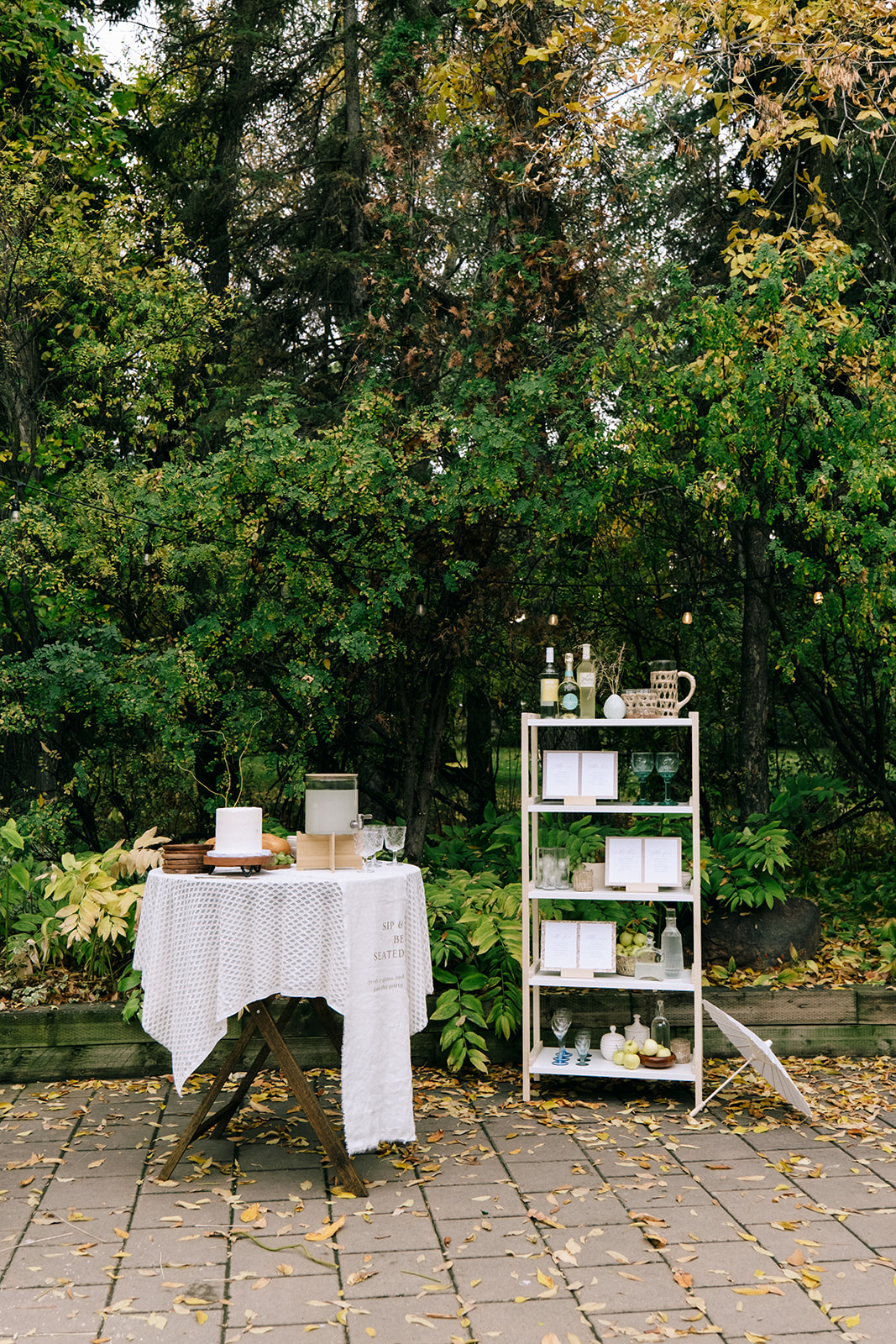 Rustic white and wood shelving display with wine bottles and vintage colored glassware for a cottage core wedding. Charming serve-yourself drink station with prosecco, vintage glassware, and a seating chart display.